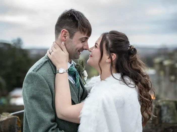 wedding photographer at Dundas Castle: a couple wearing wedding attire gazes into each other's eyes and smiles, standing outdoors with a blurred natural background. The man wears a gray suit and green tie; the woman wears a white fur shoulder wrap.