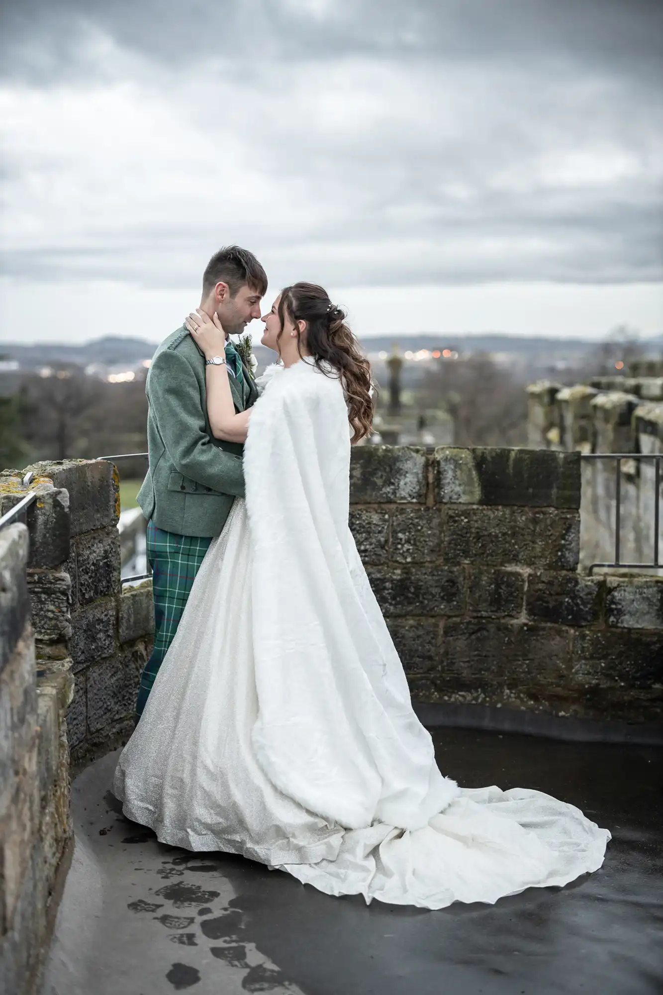 A couple dressed in formal attire stands close together on a stone walkway with city lights and a cloudy sky in the background. The woman wears a long white gown with a train, and the man wears a green jacket.