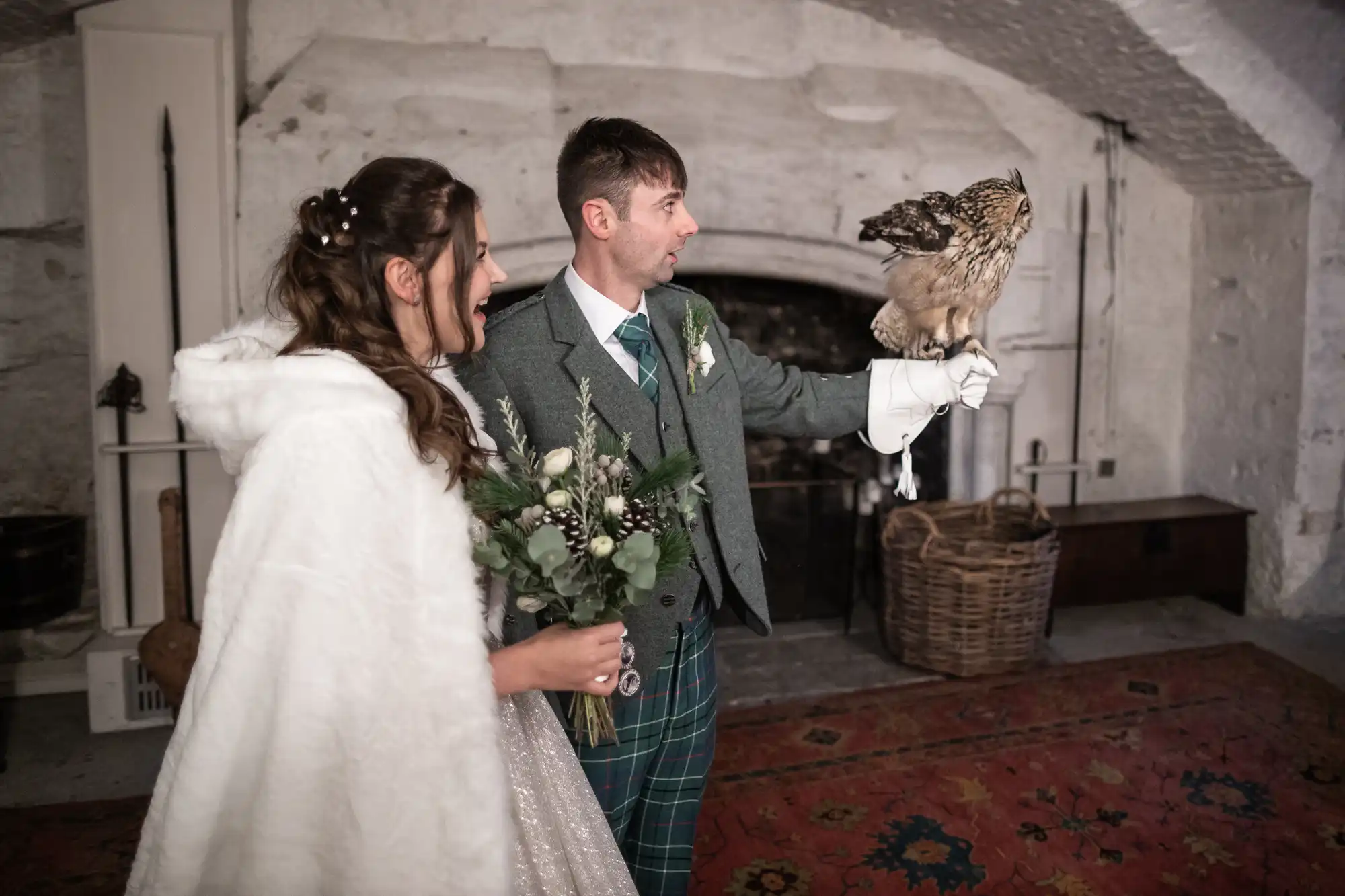 A bride and groom are indoors; the groom, holding a glove, has a bird of prey perched on his hand. The bride is holding a bouquet and looking at the bird. They are standing on a red patterned carpet.