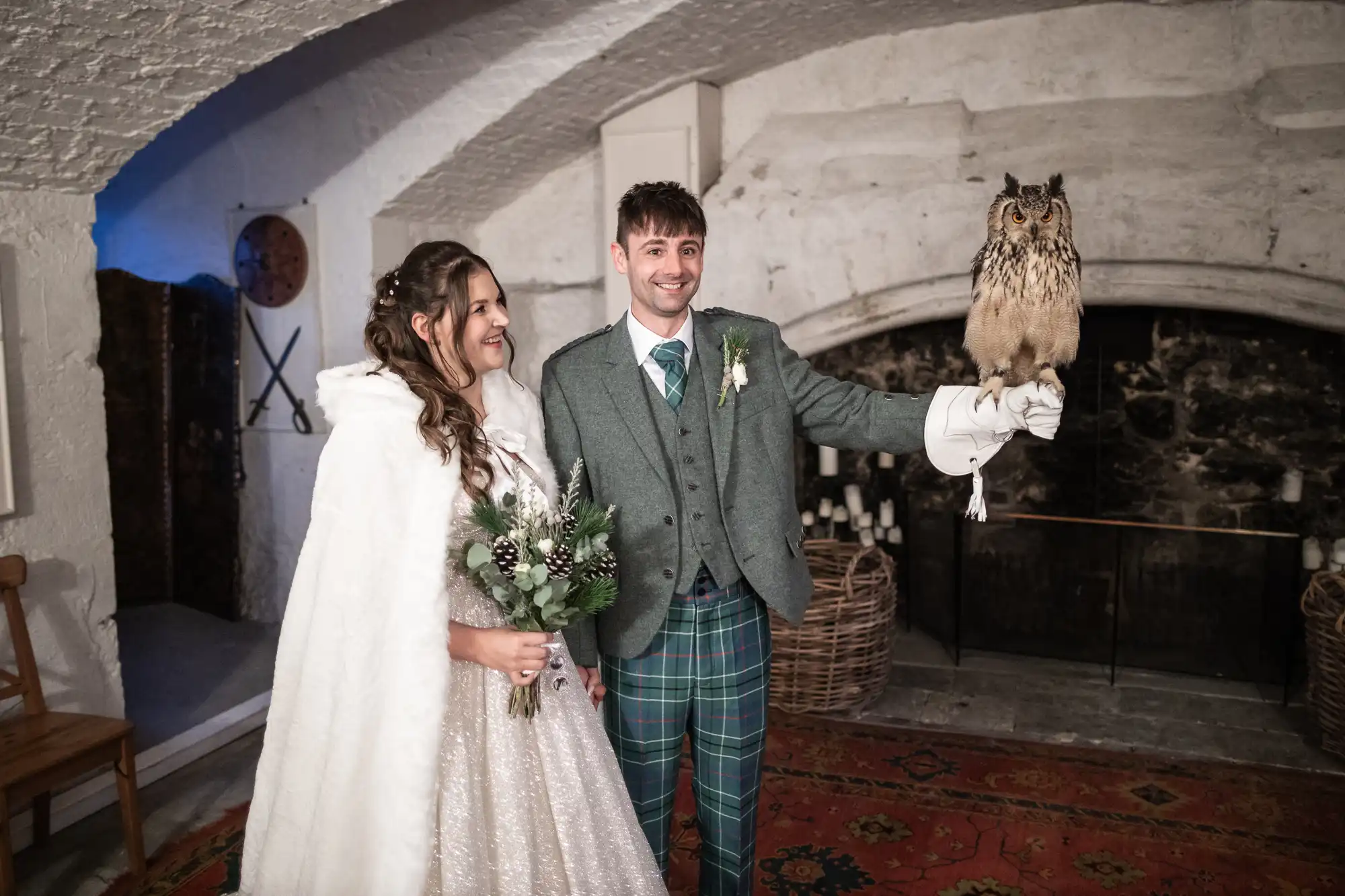 Bride and groom in wedding attire stand indoors; groom holds an owl on his gloved hand.