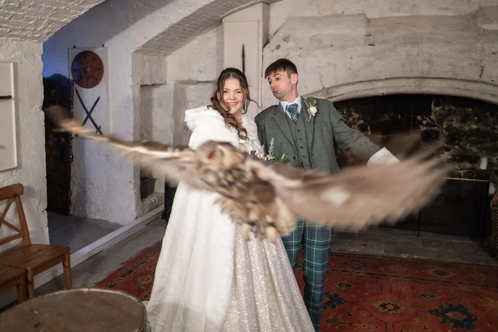 A bride and groom pose indoors while an owl flies close to the camera in front of them, captured in mid-flight.