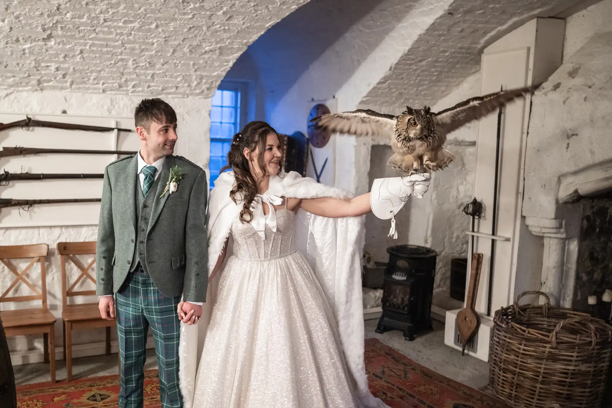 A couple in wedding attire, holding hands. The bride, wearing a white dress and cape, holds an owl on her gloved hand inside a rustic, white-walled room. The groom sports a green suit and checkered pants.