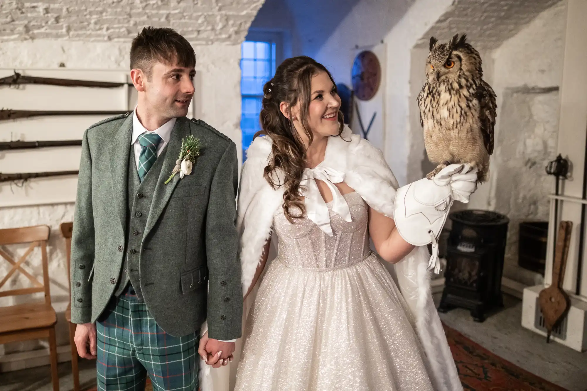 A bride in a white dress and cape holds an owl on her gloved hand, while standing next to a groom in a green suit and plaid trousers. They are indoors with weapons displayed on the wall behind them.