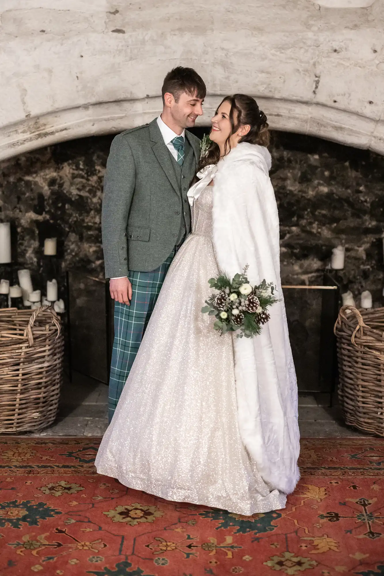 A bride and groom dressed in wedding attire stand close, smiling at each other. The bride is in a white gown and cape, holding a bouquet. The groom wears a green jacket with a plaid kilt.