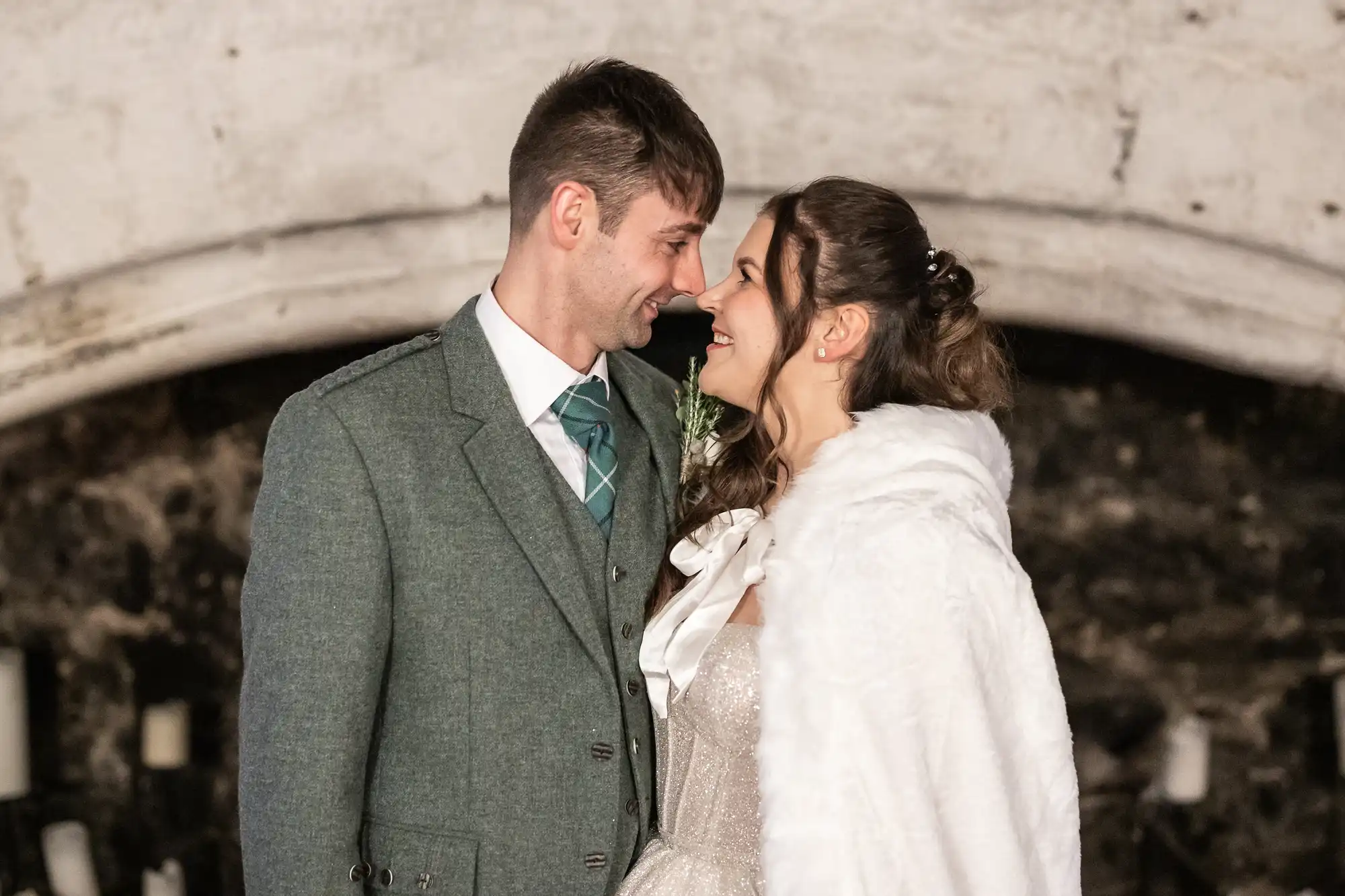 A couple in formal attire, with the man in a green suit and the woman in a white dress and shawl, face each other smiling against a stone wall background.