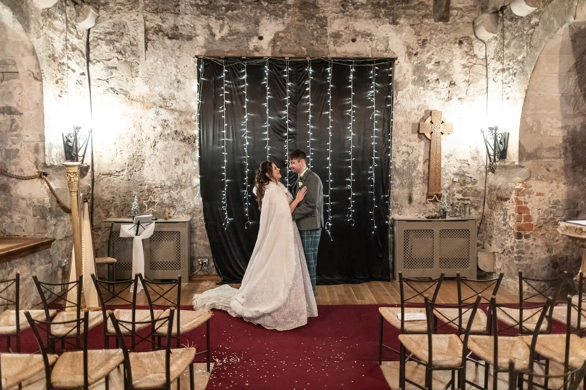 A couple stands under string lights, facing each other in a rustic stone-walled chapel with a red carpet and empty chairs. The bride wears a white gown and the groom a suit. A cross is on the wall behind them.