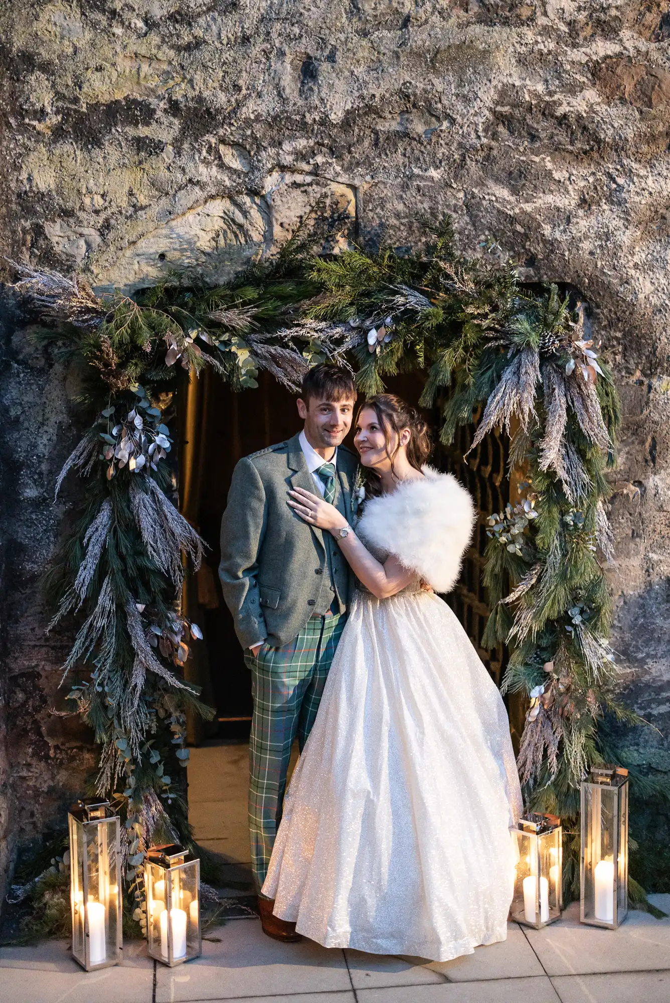 A couple stands in a rustic archway adorned with greenery and candles. The man is in a tweed suit and plaid trousers, and the woman is in a white gown with a fur stole.