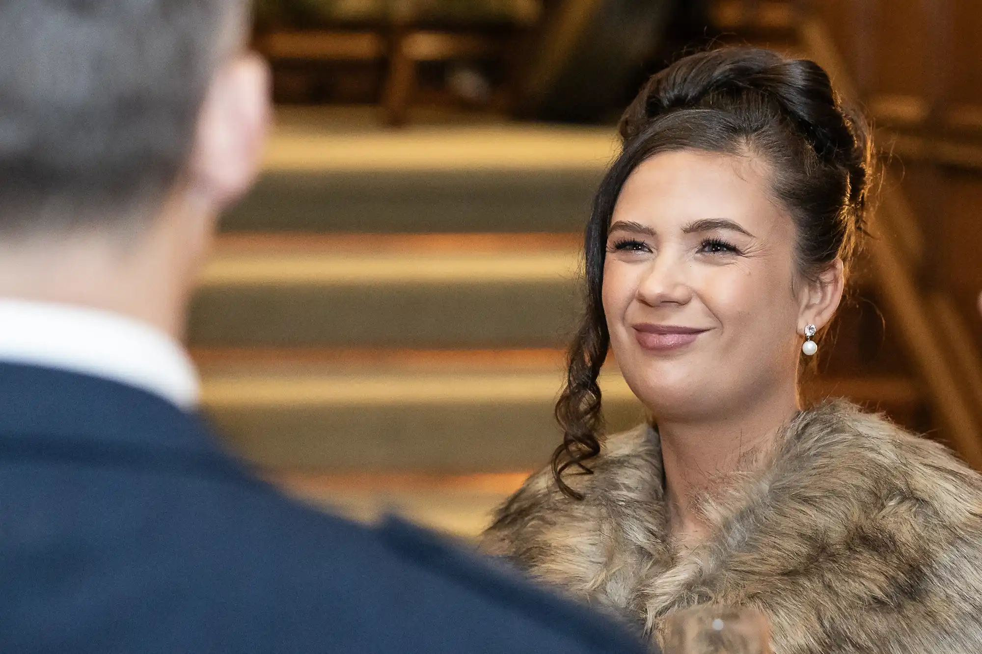 A woman with dark hair in an updo, wearing pearl earrings and a fur stole, smiles at a man with short hair in a suit. They appear to be in a warmly lit setting with stairs in the background.