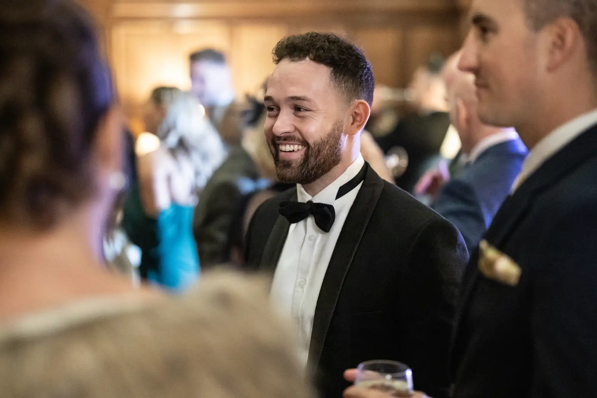 A man in a black tuxedo smiles at an event while holding a drink, surrounded by other attendees.