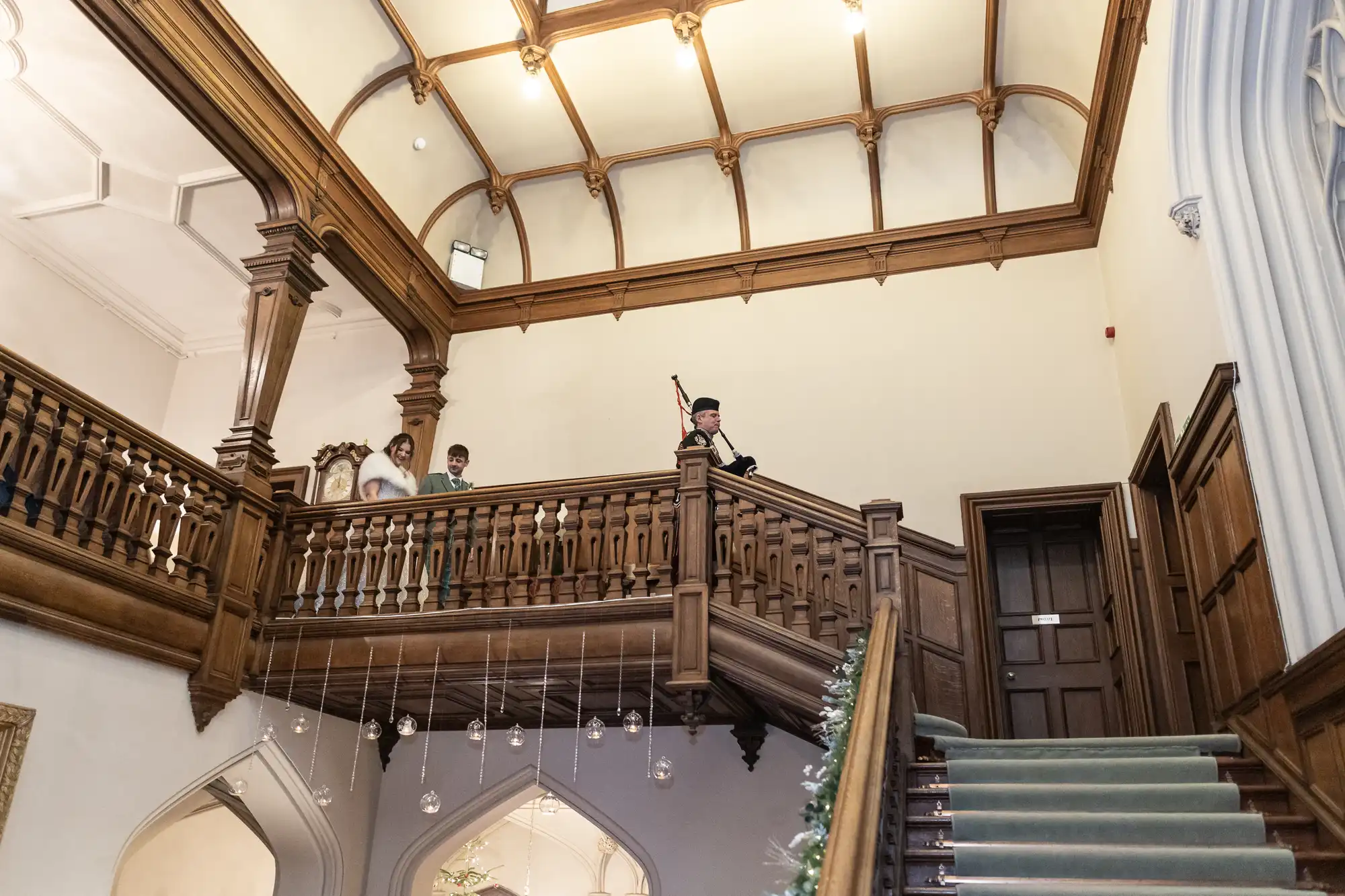 A bride and groom stand on a wooden balcony with a bagpiper in traditional attire at the top of a staircase in a historic building with arched doorways and a high, wood-paneled ceiling.