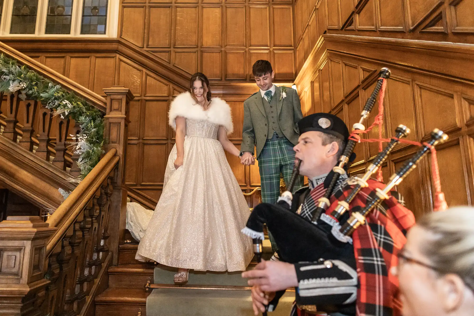 A couple descends a wooden staircase, the woman wearing a white gown and fur stole, the man in a green suit, while a bagpiper in traditional attire plays on the stairs below them.