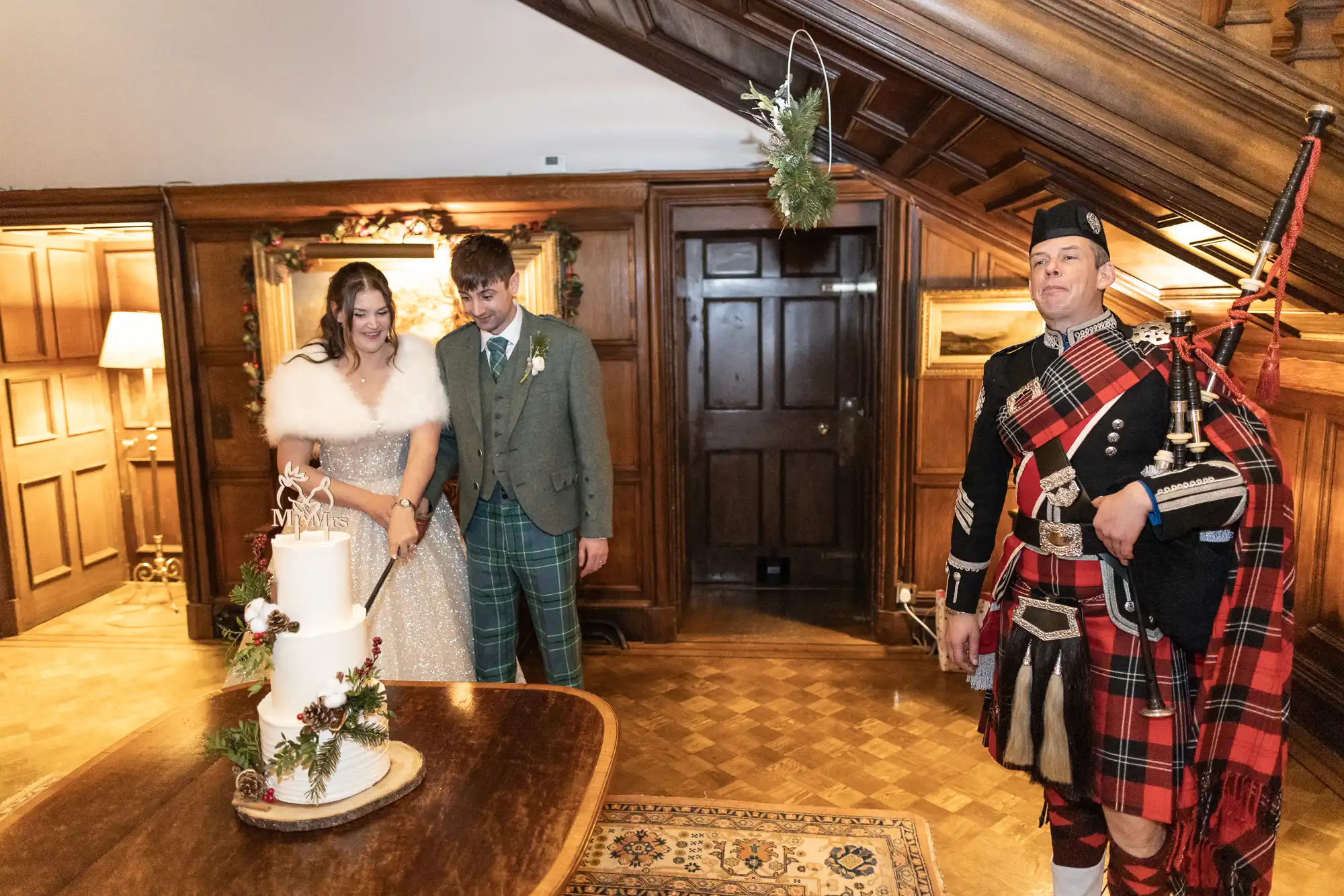 A couple stands by a decorated wedding cake with a bagpiper in traditional attire nearby. The setting appears to be an interior with wooden paneling and a staircase.