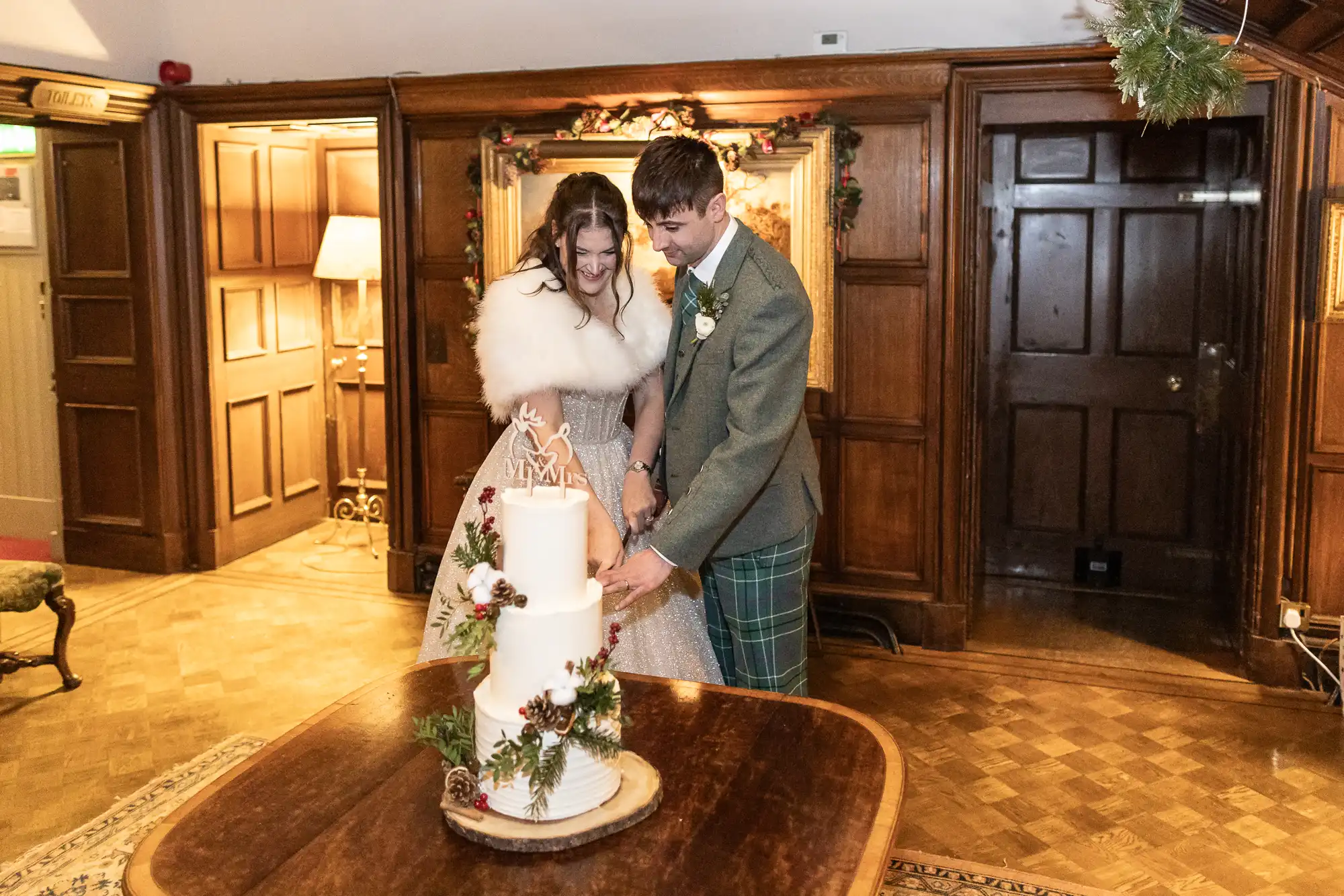 A bride and groom cut a white, three-tiered wedding cake adorned with greenery and red berries in a wood-paneled room. The bride wears a fur wrap and the groom is in a suit with plaid trousers.