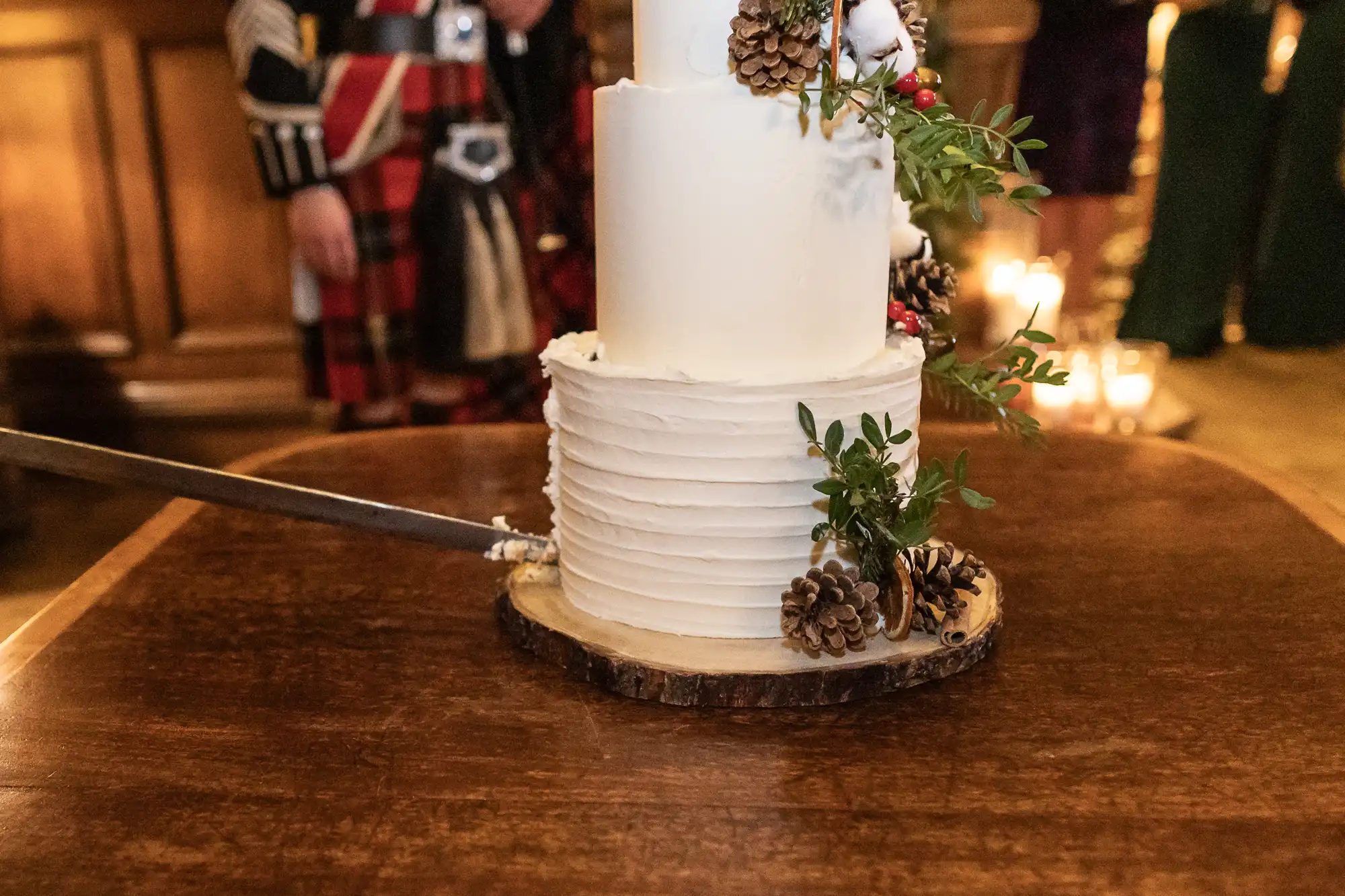 A white, three-tiered cake decorated with pine cones and greenery, resting on a wooden slice. A knife is partially inserted into the side of the cake.