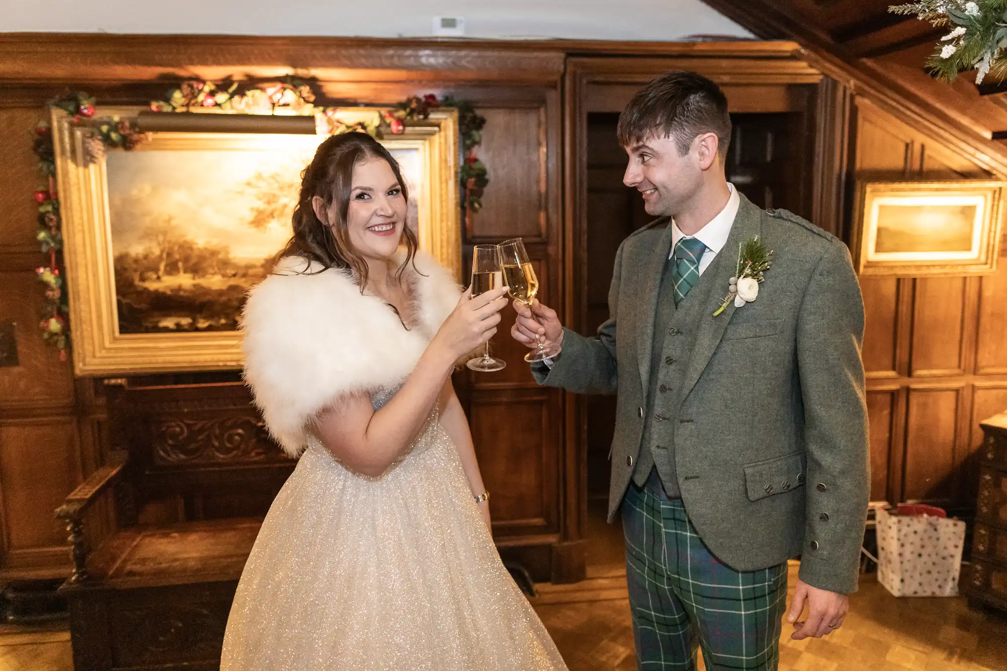 A bride in a white gown with a fur shawl and a groom in a green suit with a kilt cheer with champagne glasses in a wood-paneled room.