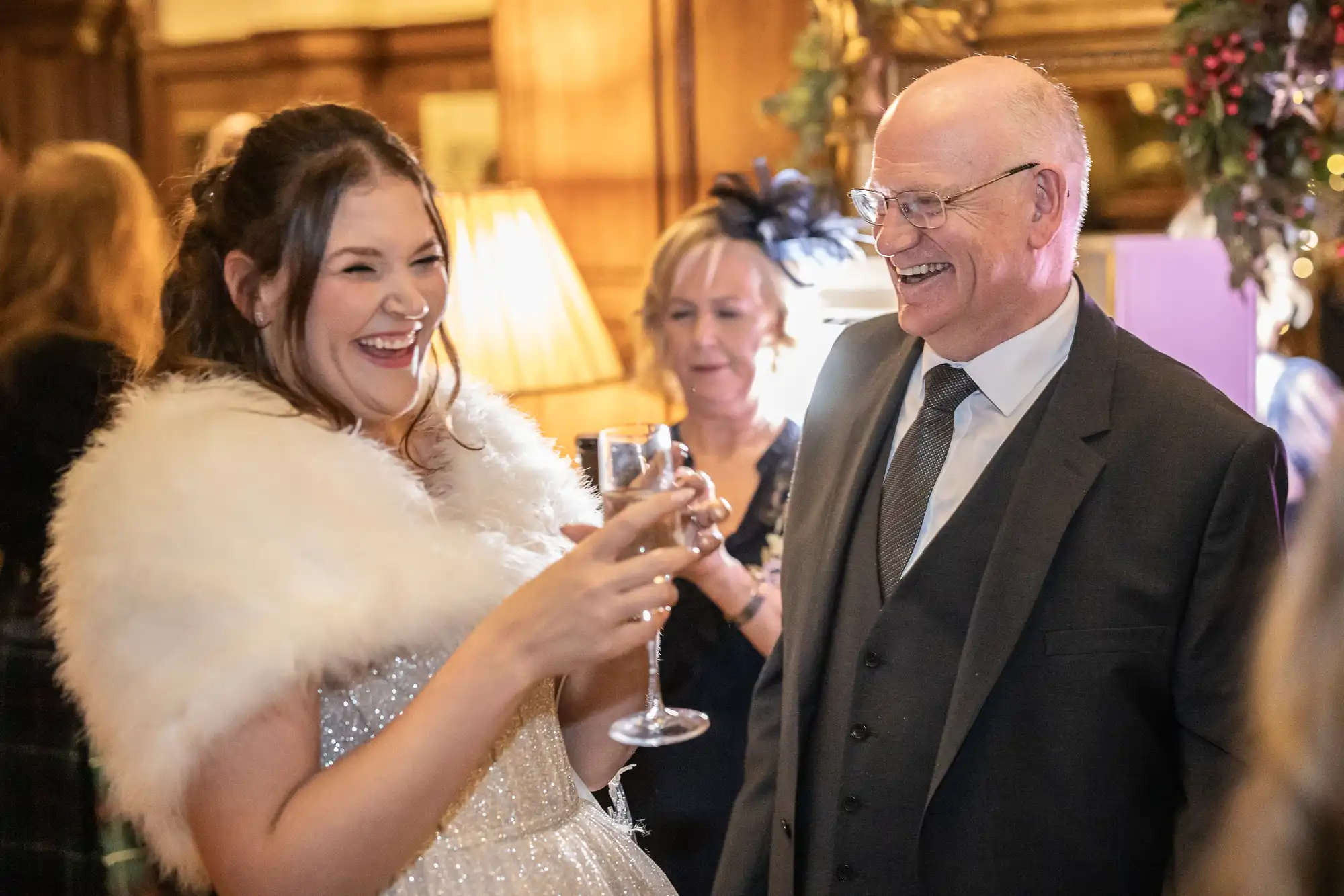 A young woman in a white dress and fur shawl laughs while holding a wine glass, beside an older man in a suit and tie, both smiling in a warmly lit room.