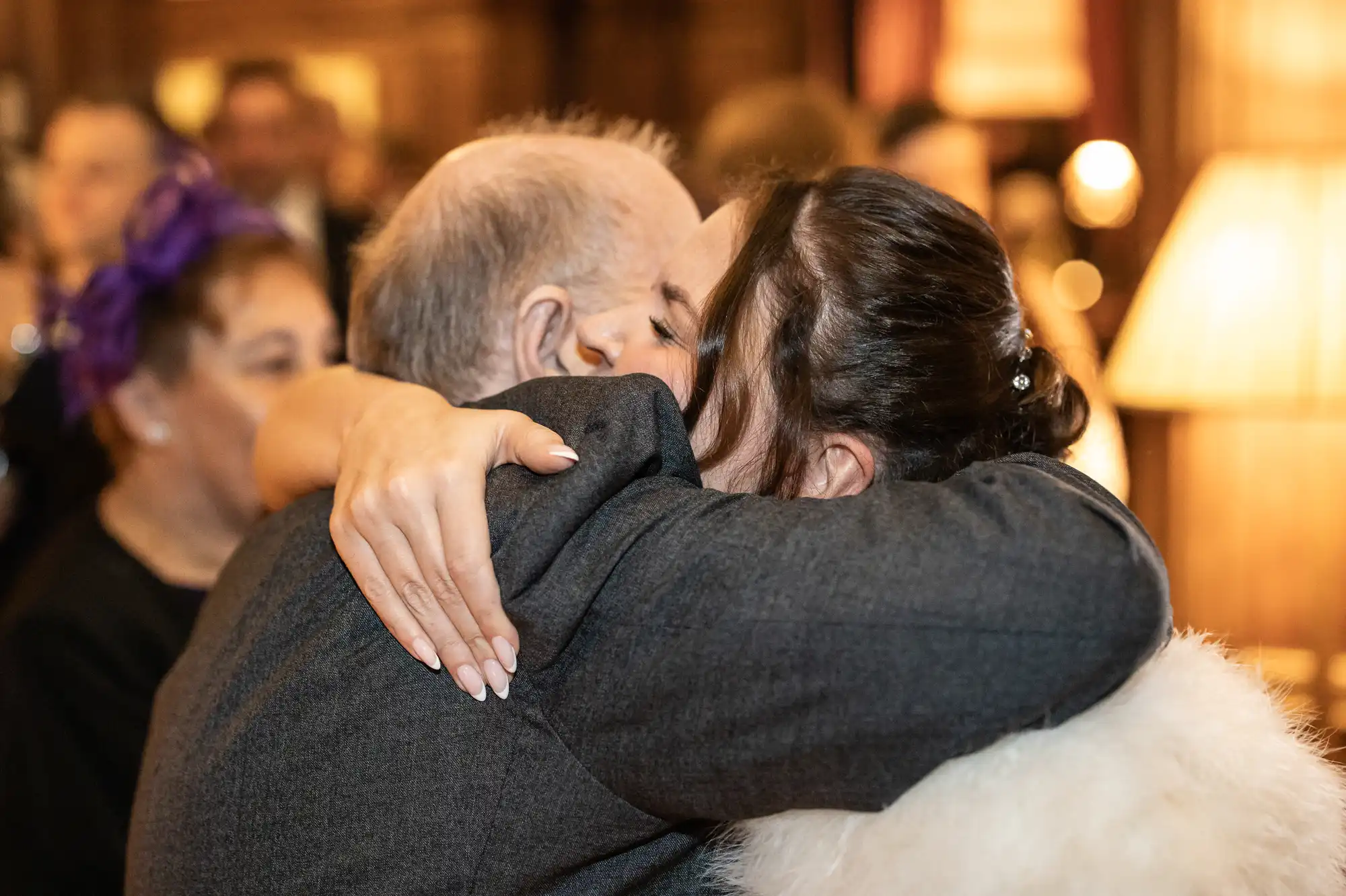 An older man and a younger woman embrace at a social gathering, with their faces not visible. People and soft lighting are seen in the background.