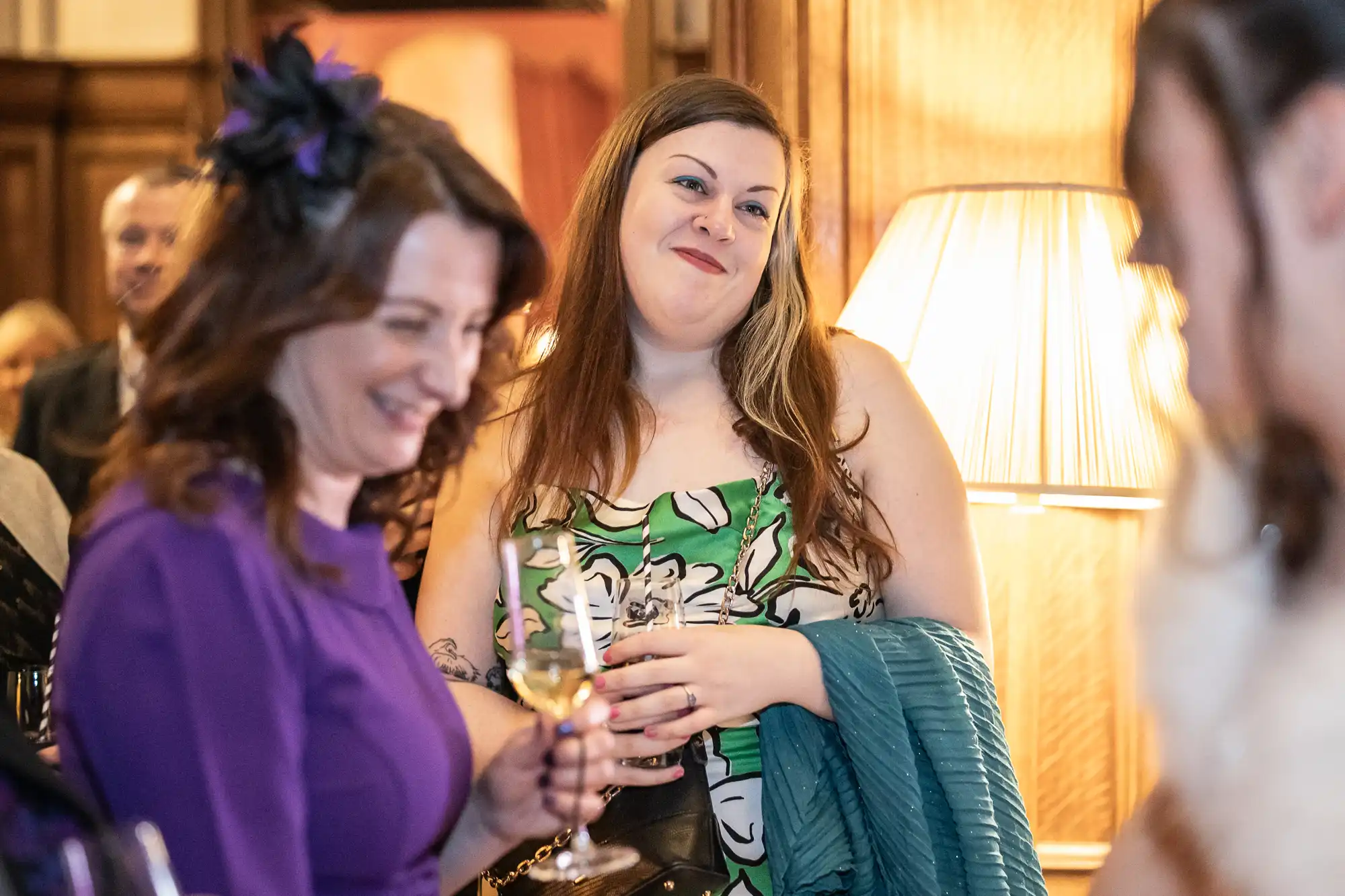 Two women stand near a lit lamp at a social event, holding drinks and engaged in conversation. One woman is smiling and wearing a purple outfit, while the other wears a green patterned dress and holds a jacket.