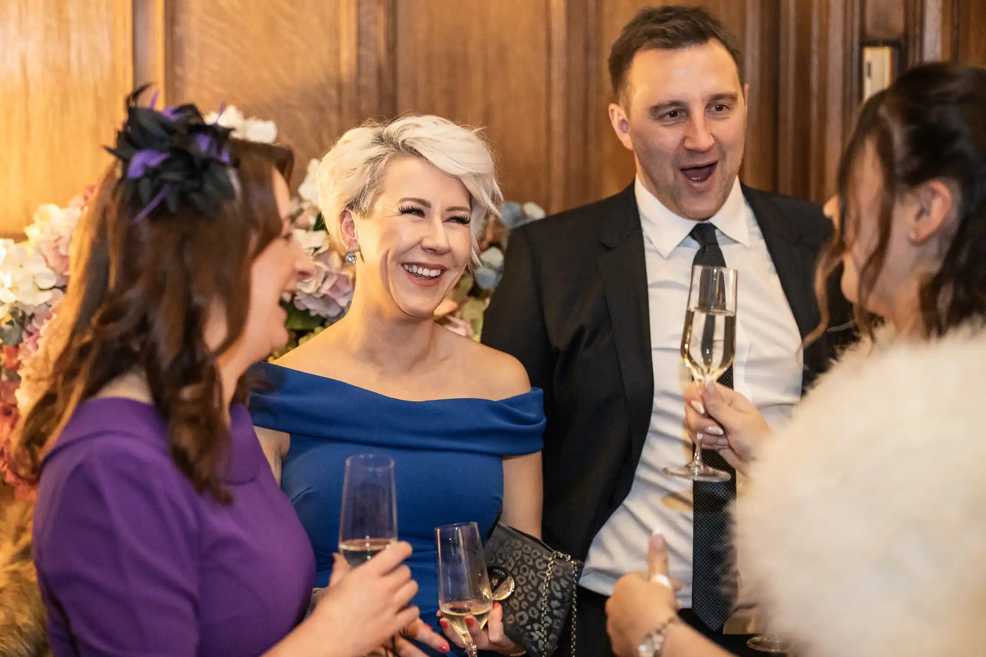 Four people dressed in formal attire are standing together, holding glasses of champagne, and smiling while engaged in conversation at an indoor event.