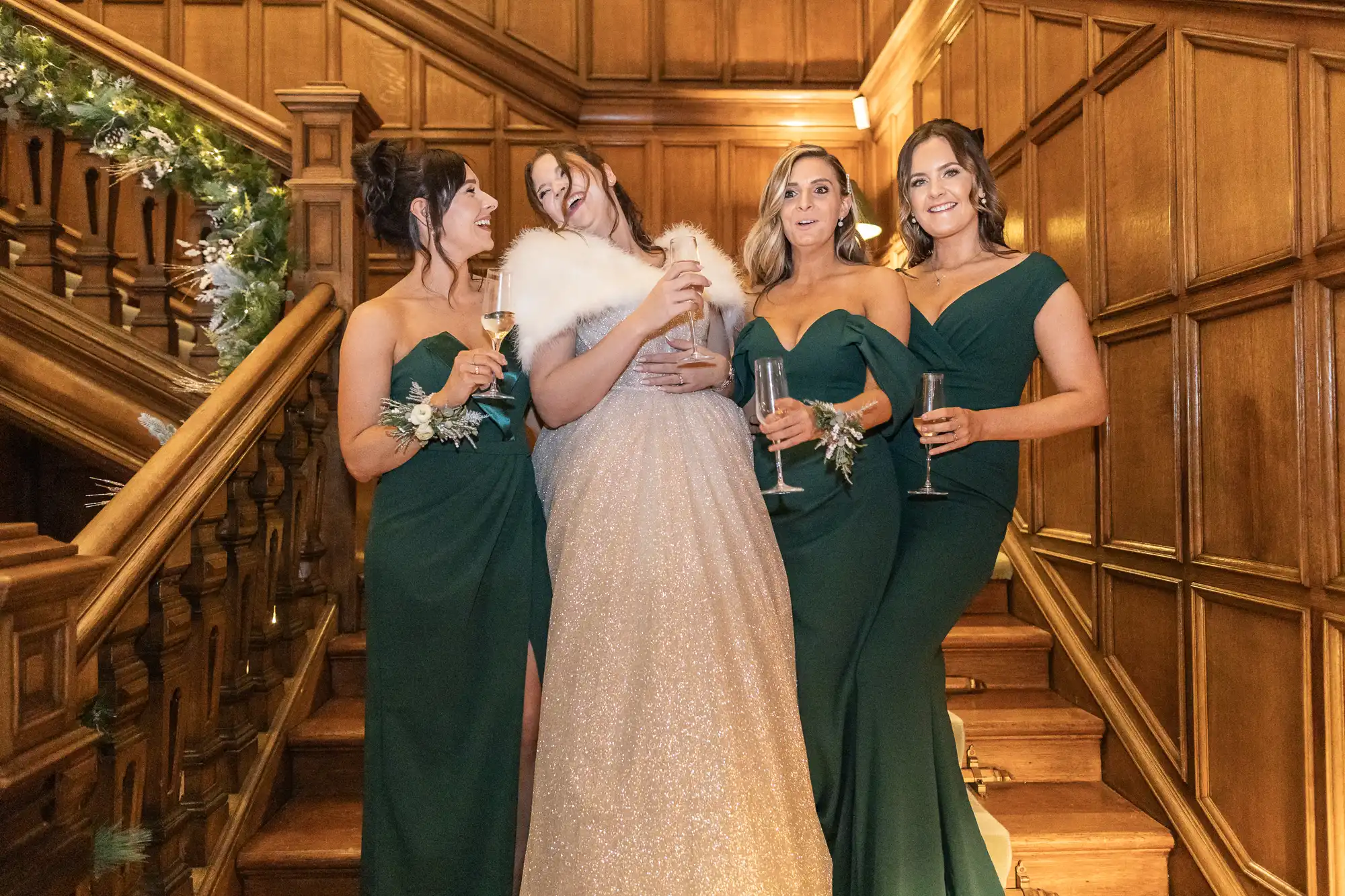 A bride in a sparkling dress and white shawl stands on a wooden staircase with three bridesmaids in green dresses, all holding champagne glasses.