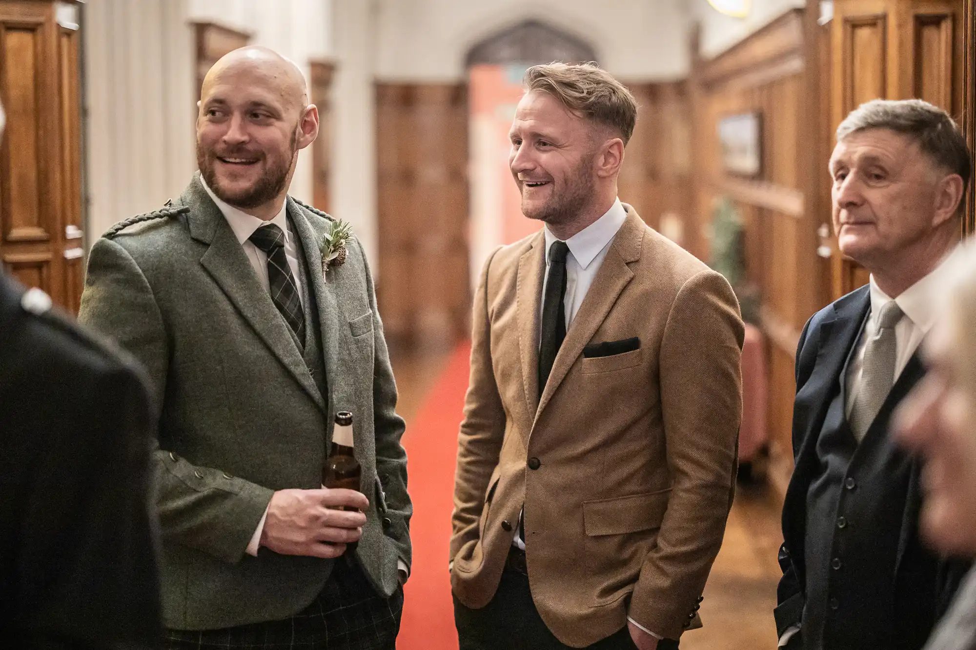 Three men, dressed in suits and ties, stand closely together, conversing and smiling in a warmly lit indoor setting. One holds a bottle, possibly at a formal event or gathering.