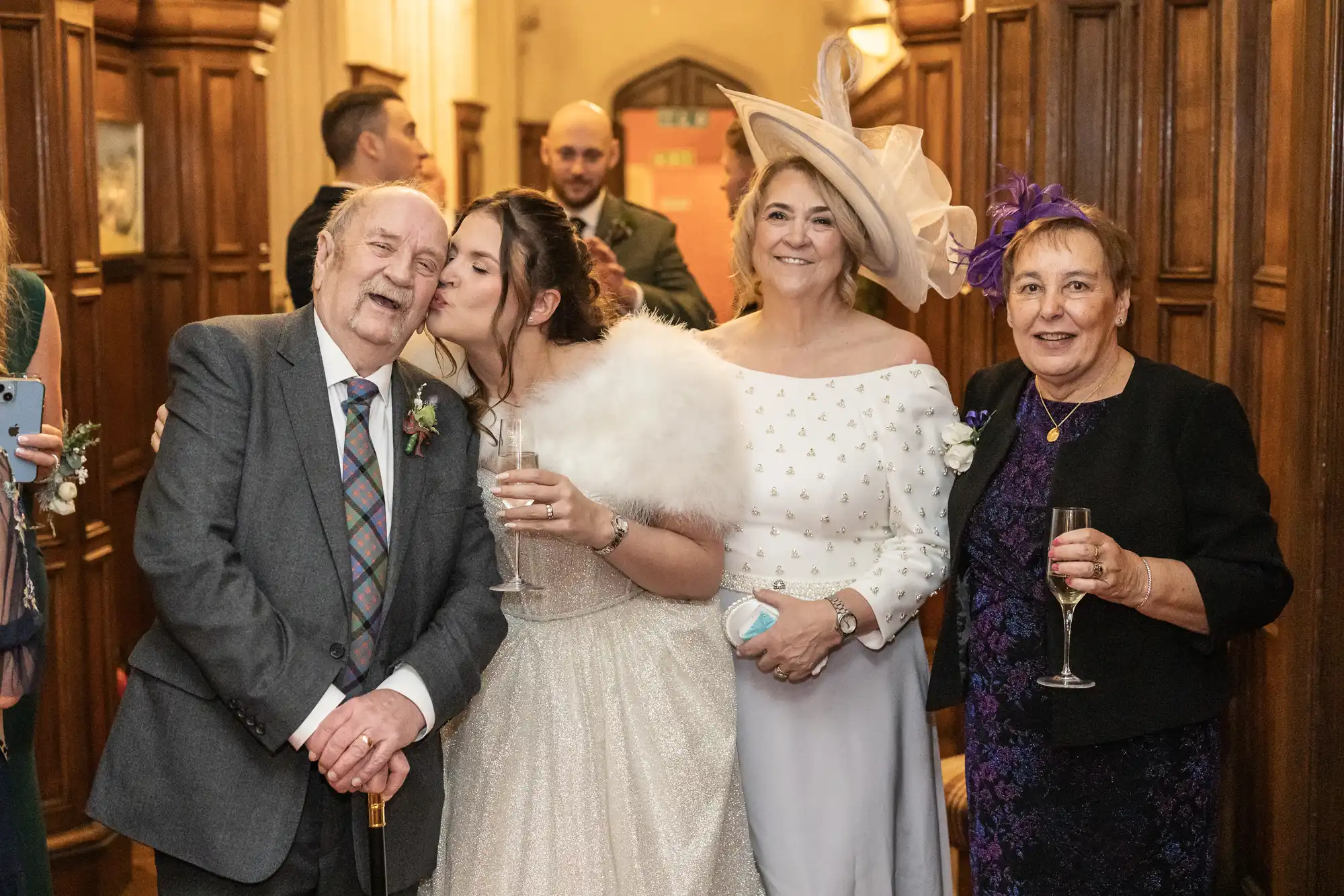 A woman in a white fur wrap kisses an elderly man on the cheek while holding a drink. Another woman in white with a large hat and a woman in black with a purple fascinator stand next to them, smiling.