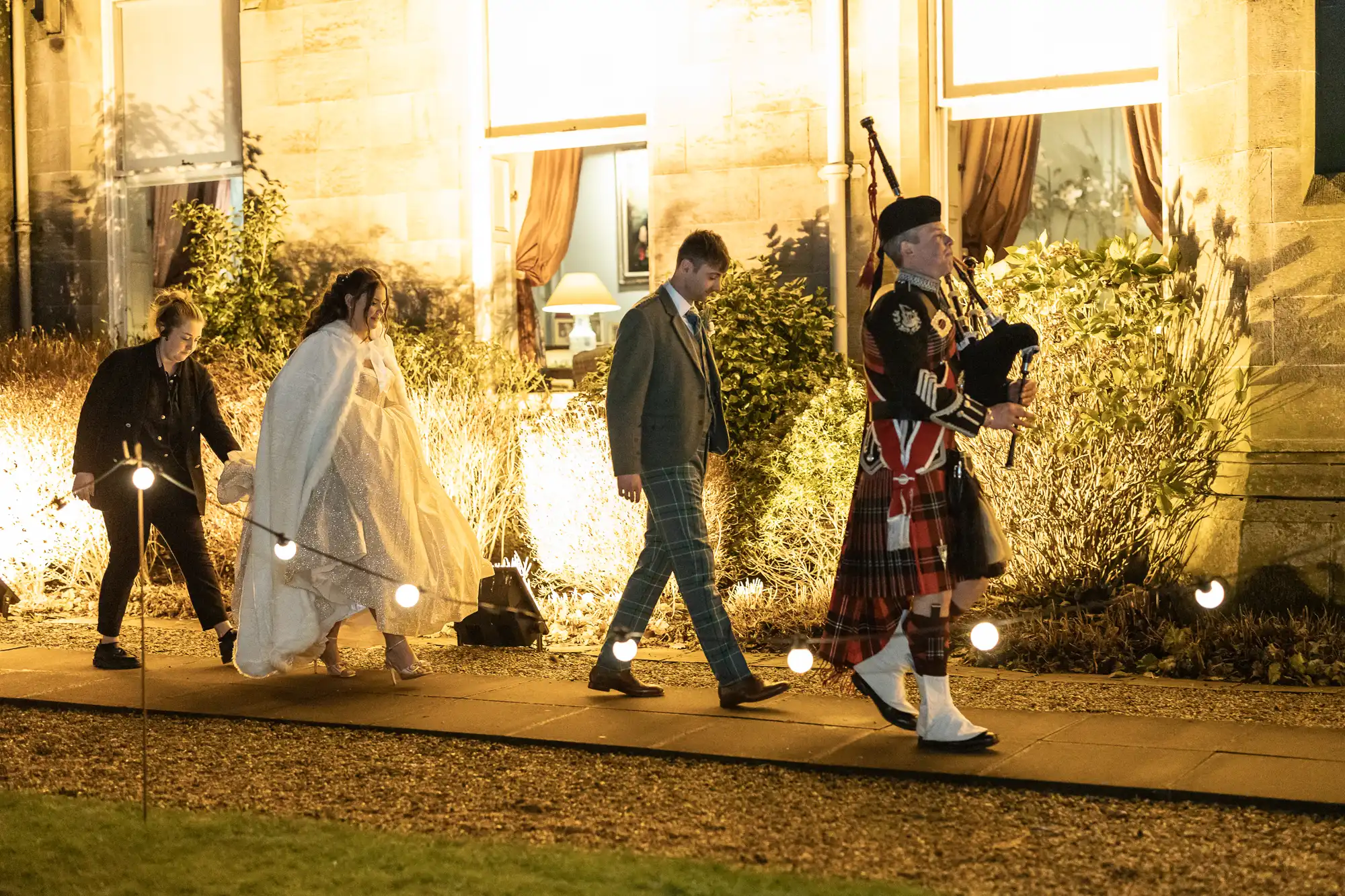 A bride in a white dress and cloak, followed by a groom in a suit, a guest, and a bagpiper in traditional attire, walk along a lit path outside a building at night.