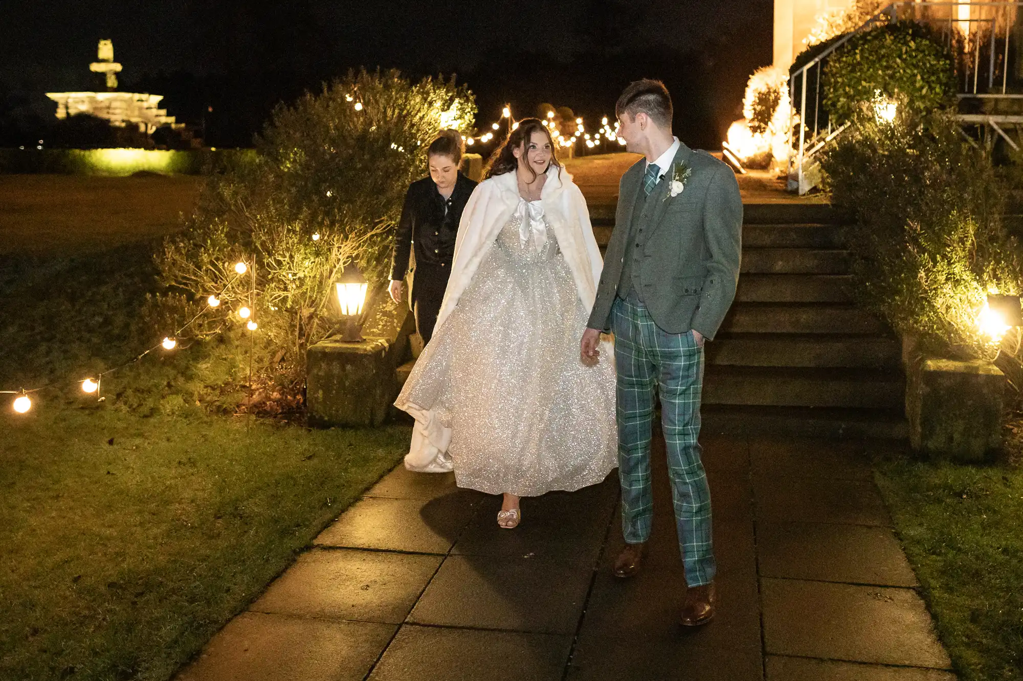 A bride and groom, dressed in formal attire, walk outside along a lit path at night, accompanied by another person. The bride wears a white dress and cape, while the groom wears a suit with plaid pants.