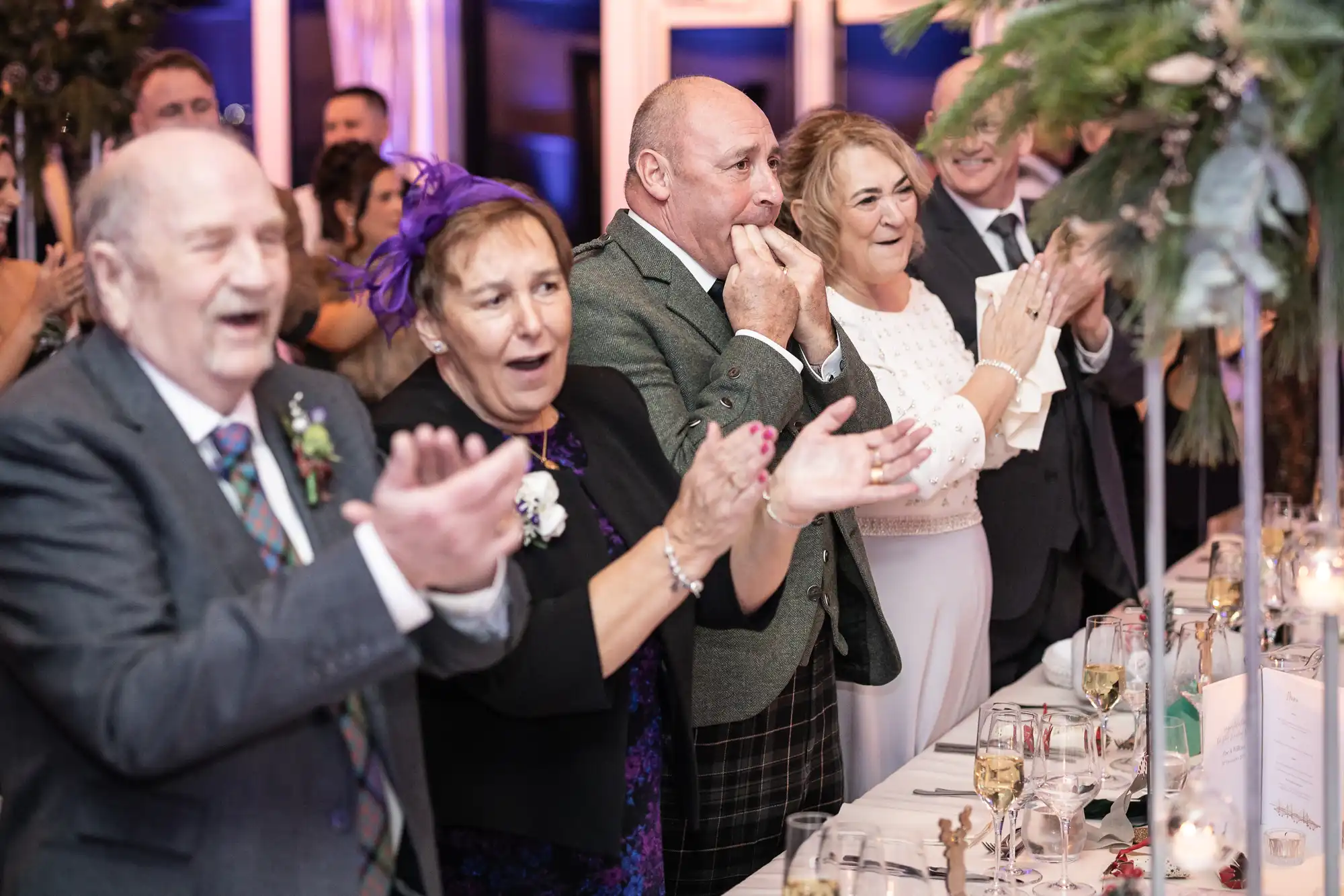 A group of people dressed in formal attire stand and clap enthusiastically at a wedding reception. They appear to be smiling and celebrating.