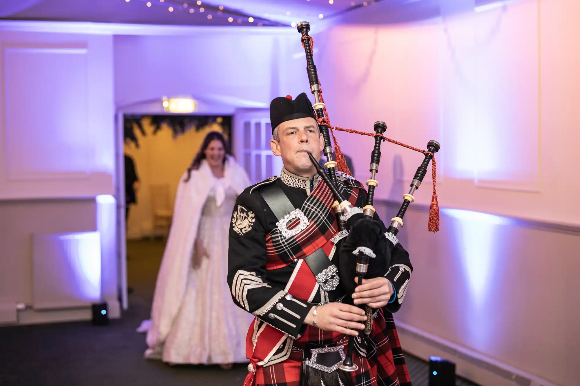 A man dressed in traditional Scottish attire plays the bagpipes as he leads a procession. A woman in a white gown follows behind him. The room is illuminated with purple and blue lights.