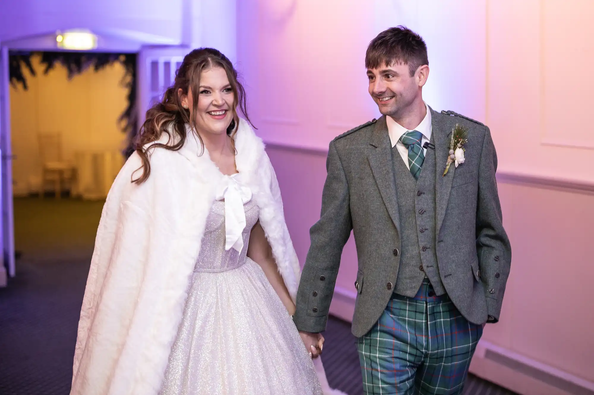 A bride in a white dress and fur cape holds hands with a groom in a green plaid kilt and gray jacket. They are indoors with purple lighting.