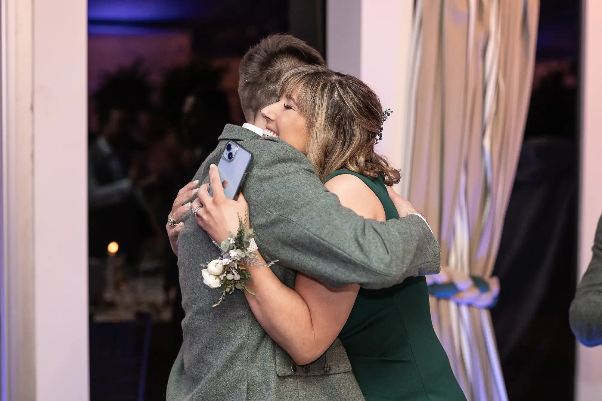 A woman in a green dress and a man in a suit embrace. The man holds a smartphone, and the woman wears a wrist corsage. They are indoors with curtains and dim lighting in the background.