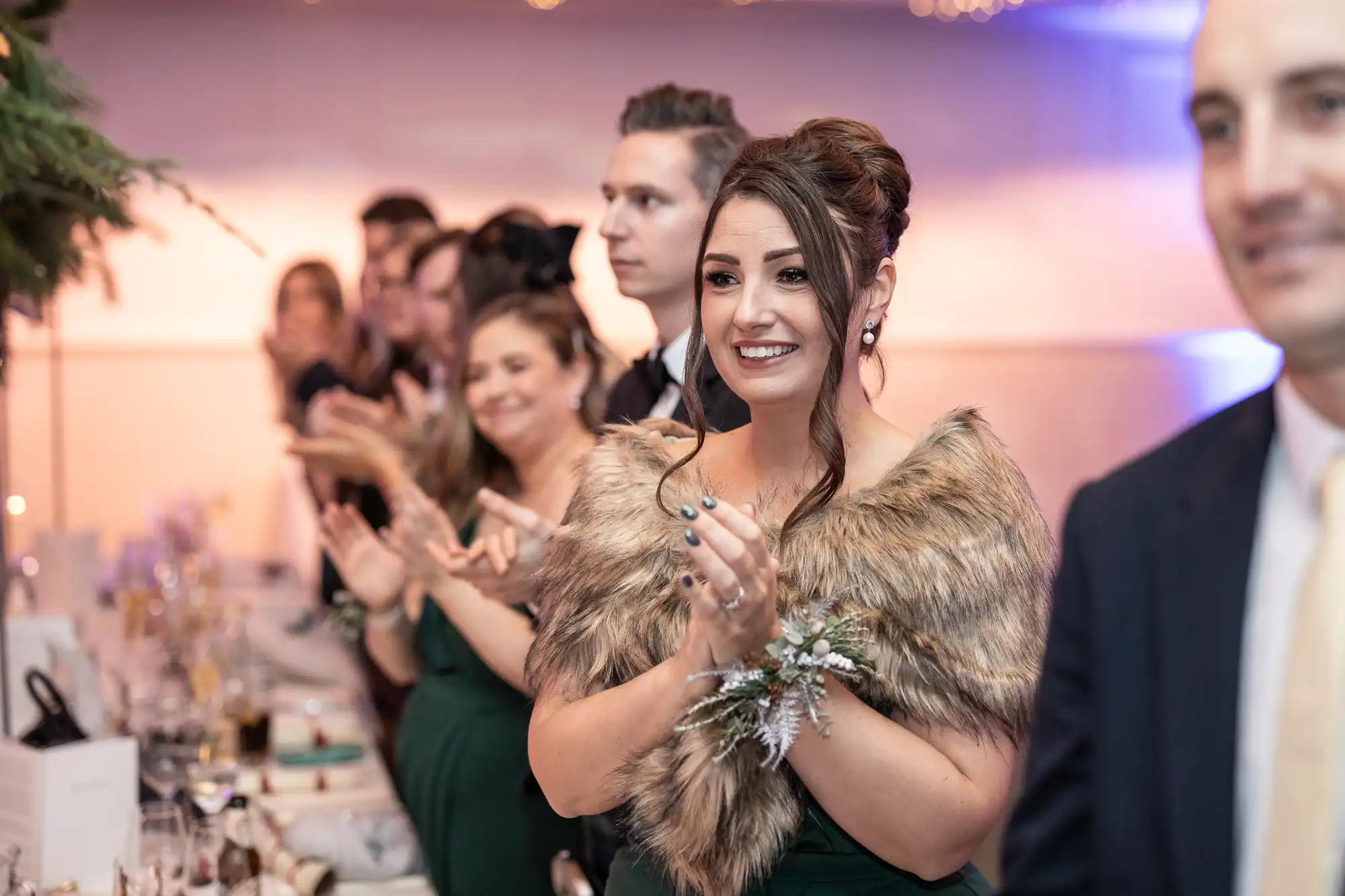 A group of people dressed formally stand in a line, applauding. The woman in the foreground wears a fur shawl and a green dress, smiling as she claps.