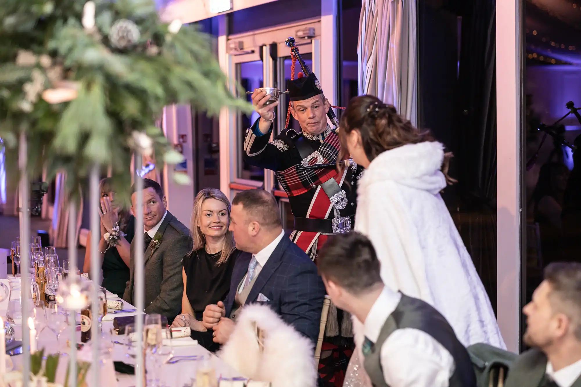 A person dressed in traditional Scottish attire raises a glass while standing at a table during a formal gathering, with attendees seated and engaging in conversation.