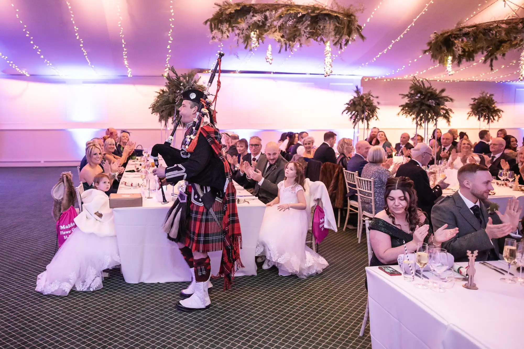 A bagpiper in traditional Scottish attire plays while walking through a wedding reception, with guests seated at tables applauding and smiling.