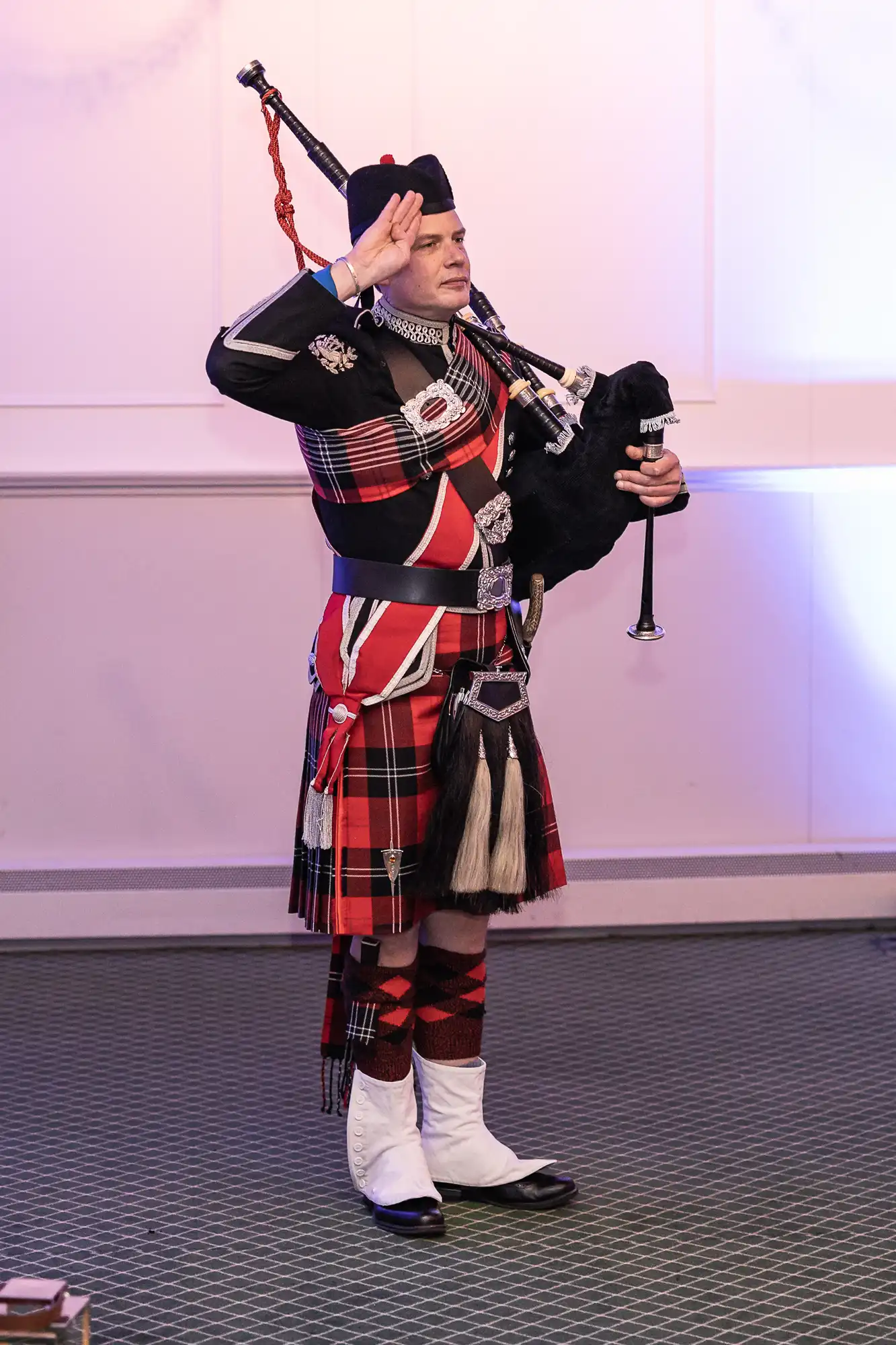 A bagpiper in a traditional Scottish outfit stands on a green carpet, saluting while holding bagpipes.