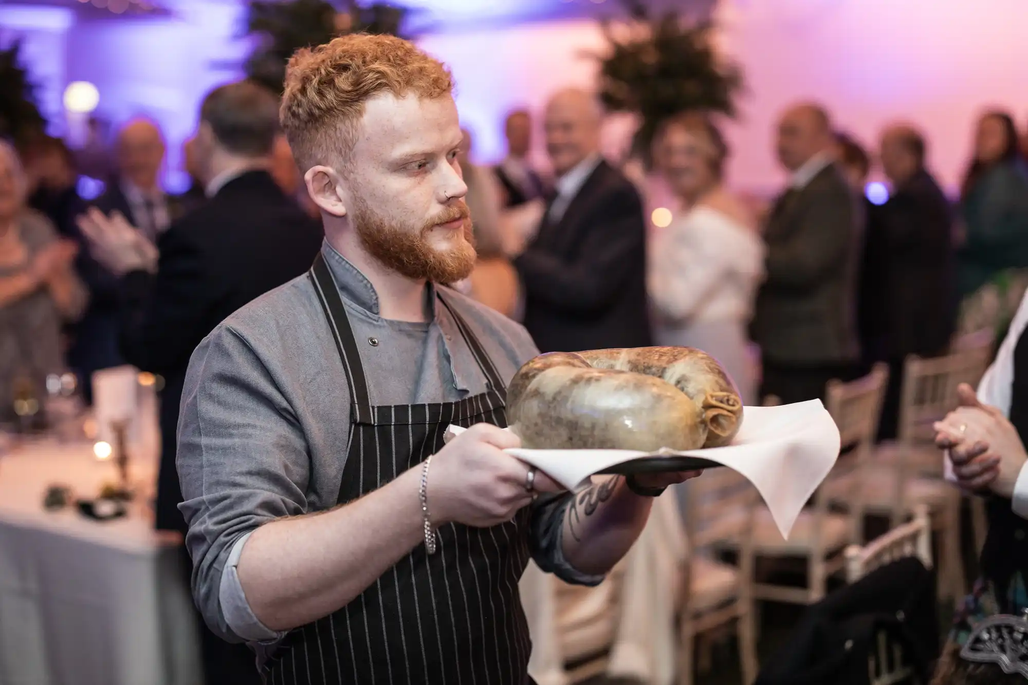 A chef with a red beard and wearing an apron holds a large dish, with an audience in the background.