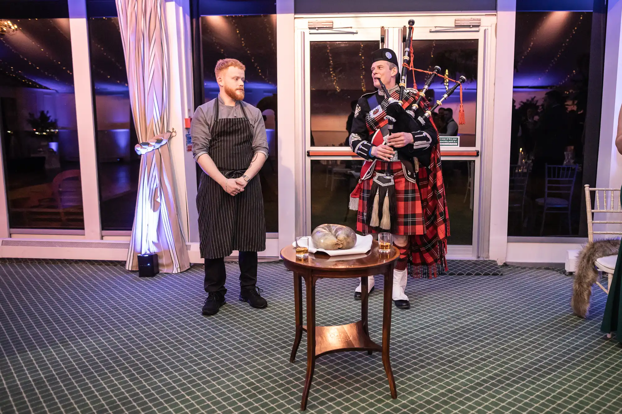 A person in a chef's uniform stands next to a bagpiper in traditional attire. A table in front holds a covered dish, with the setting in an indoor venue.