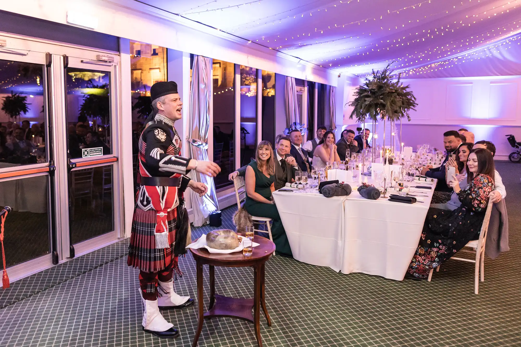 A man in traditional Scottish attire speaks to a seated audience at a formal event. The room is decorated with fairy lights and tables are set with candles and floral centerpieces.