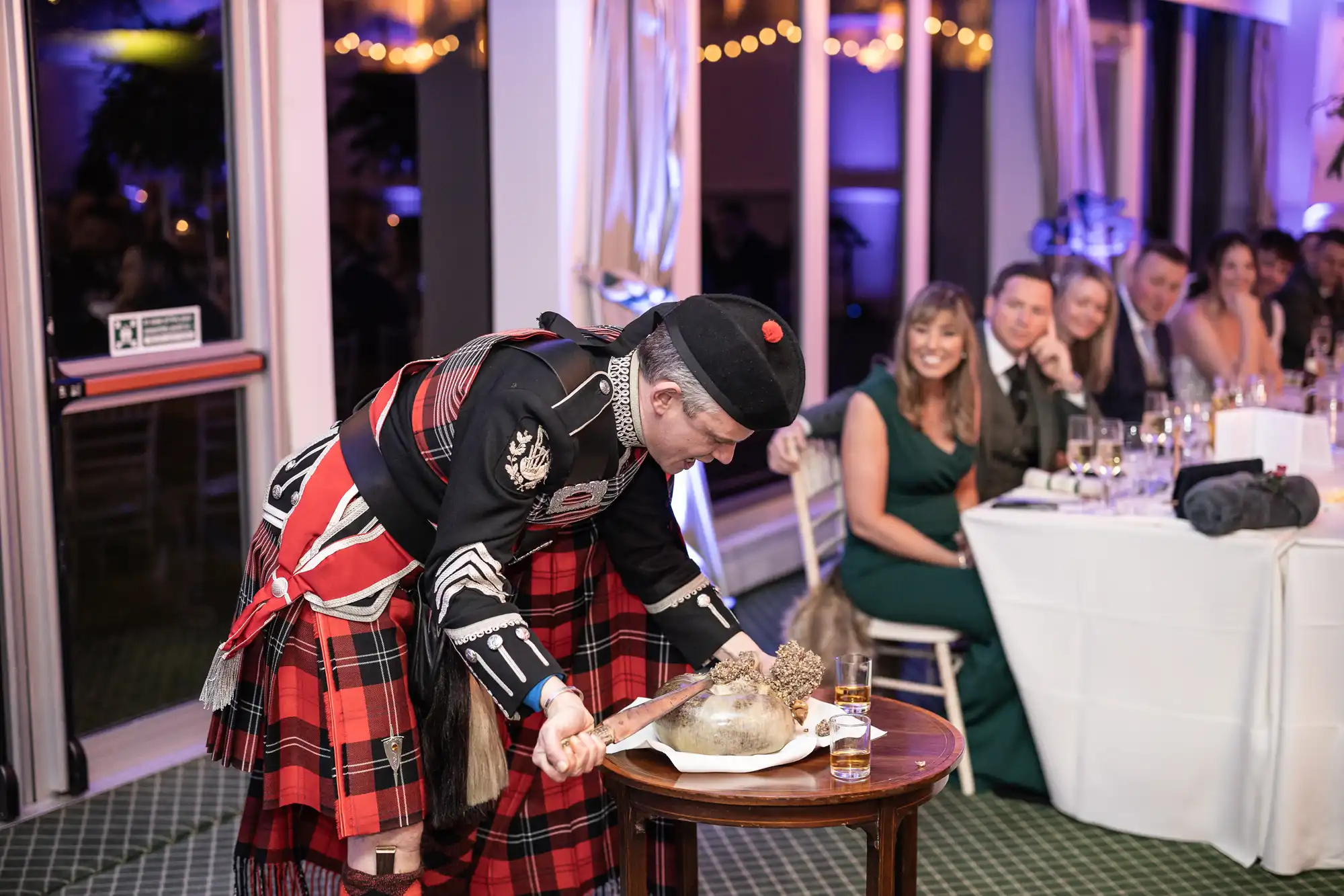 A man in traditional Scottish attire is preparing to cut a haggis on a table during an event. Guests seated nearby observe with interest.