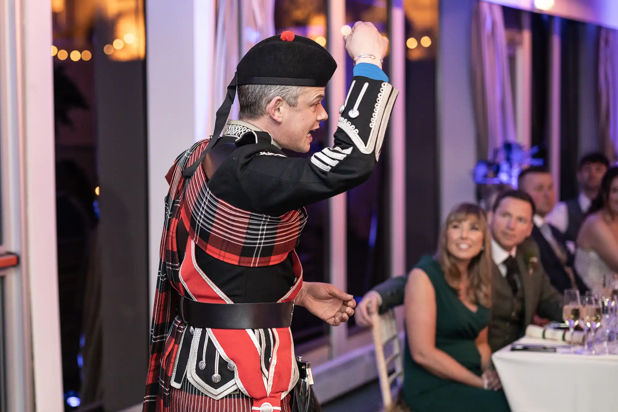 A man in traditional Scottish attire, including a tartan sash and a hat, performs with his fist raised in front of seated guests at an indoor event.