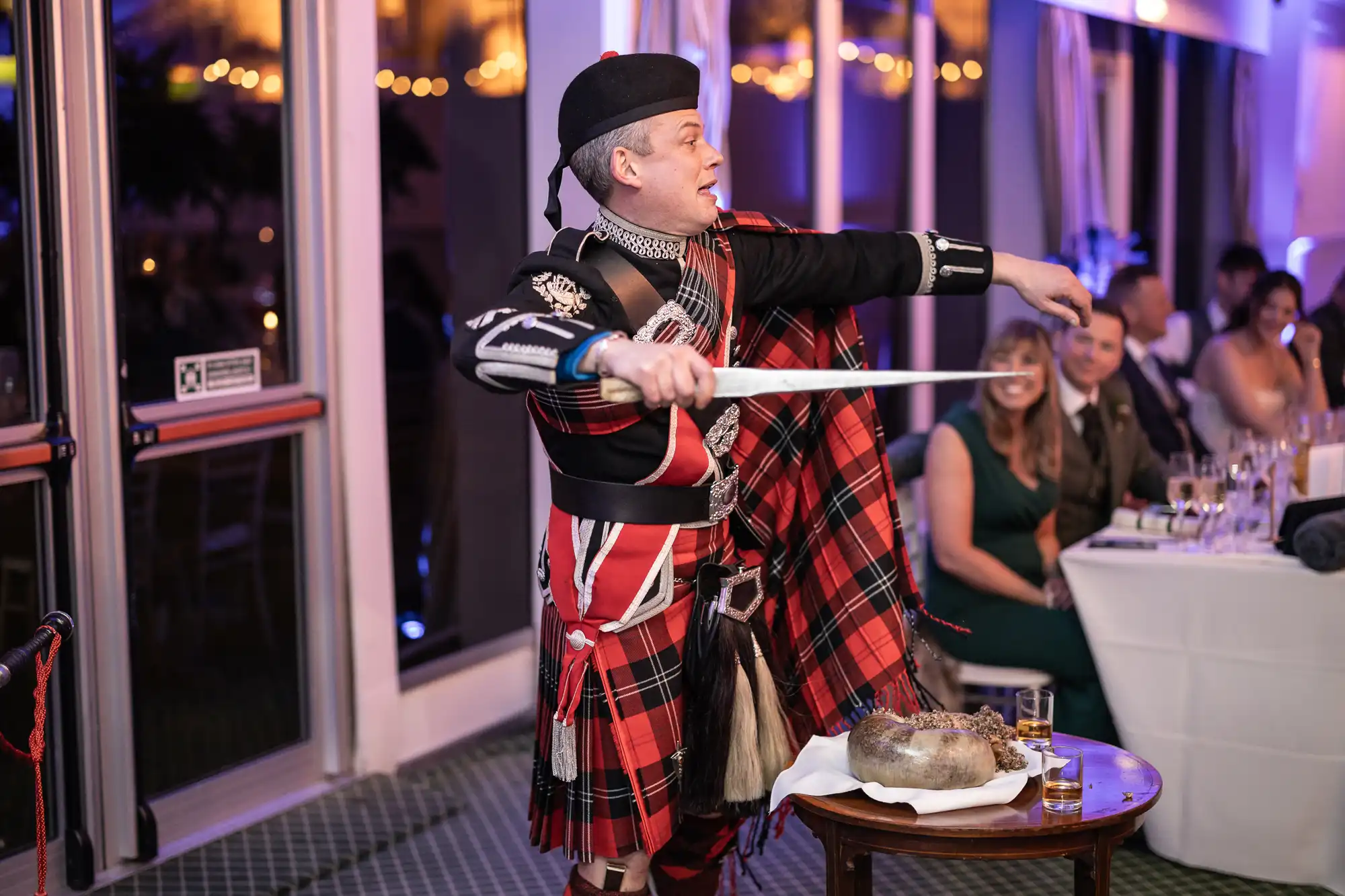 A man in traditional Scottish attire performs a haggis-cutting ceremony with a knife. He is standing next to a small table holding a haggis, with guests seated in the background.