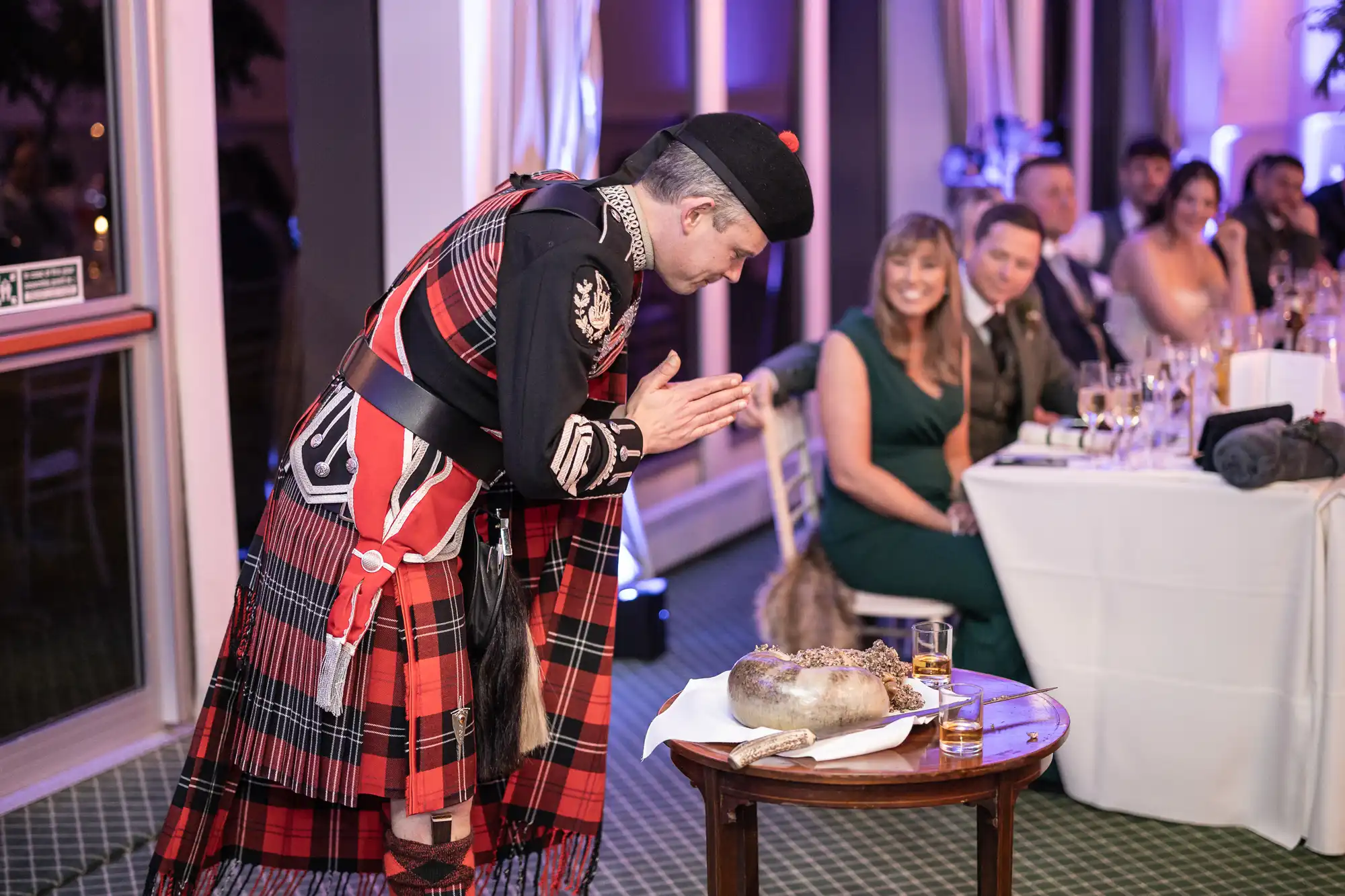 A man dressed in traditional Scottish attire bows respectfully before a haggis placed on a table. Seated guests watch and smile at the event, which appears to be a formal gathering or celebration.