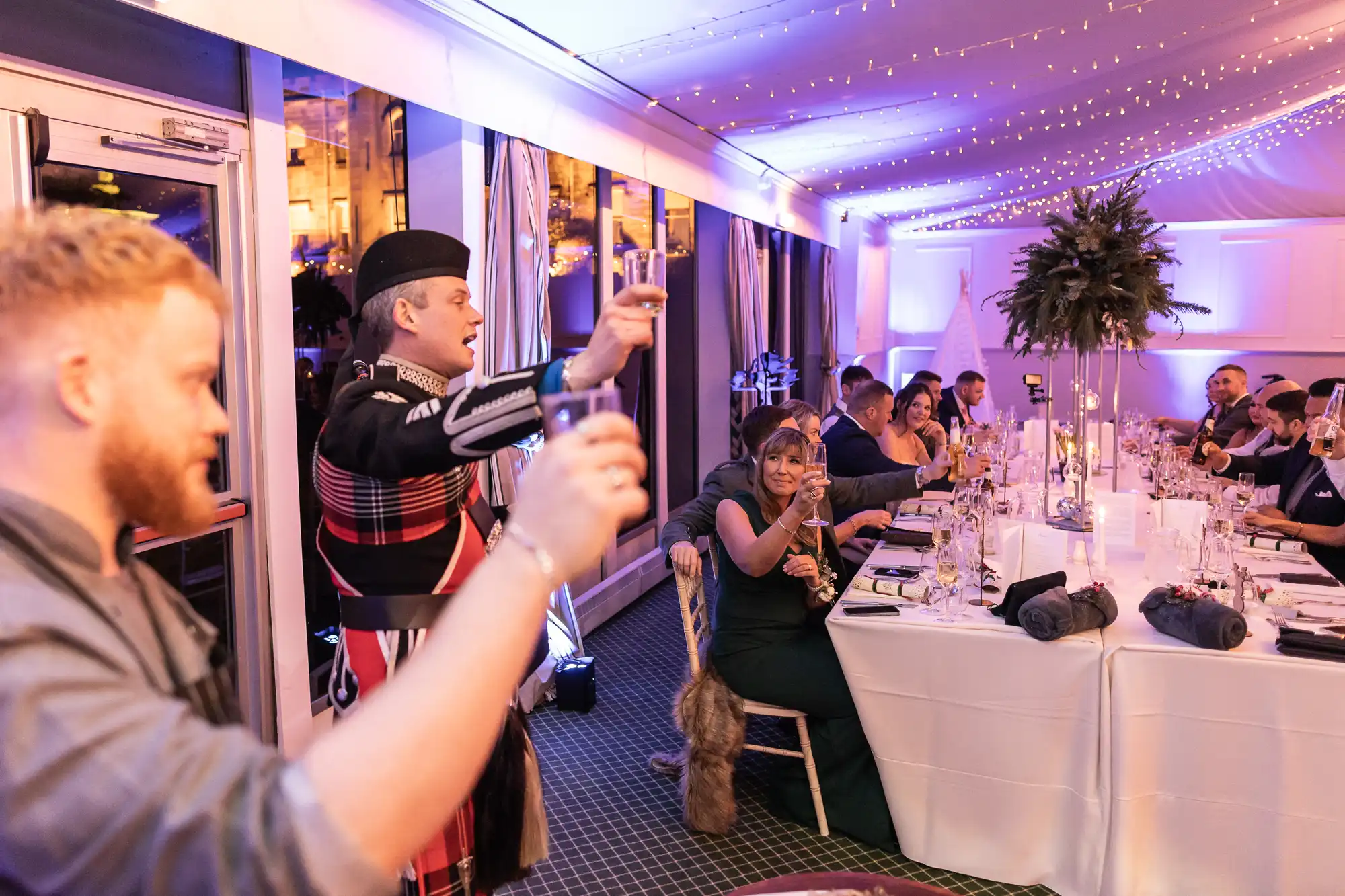 A group of people seated at a long table raise their glasses in a toast. A man in traditional Scottish attire leads the toast in a well-lit, decorated room.