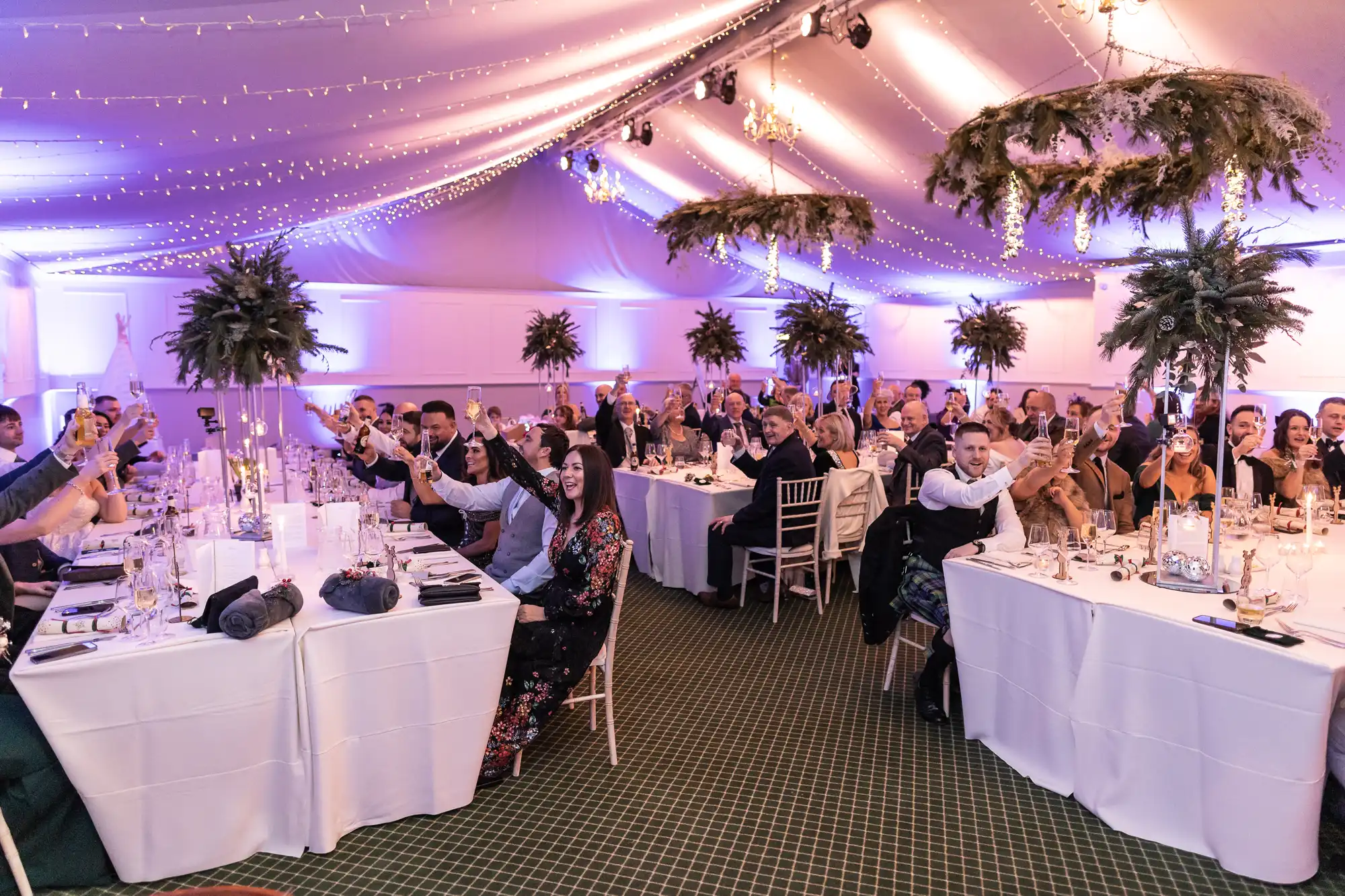 A large group of people seated at round tables inside a decorated tent raise their glasses in a toast during an event.