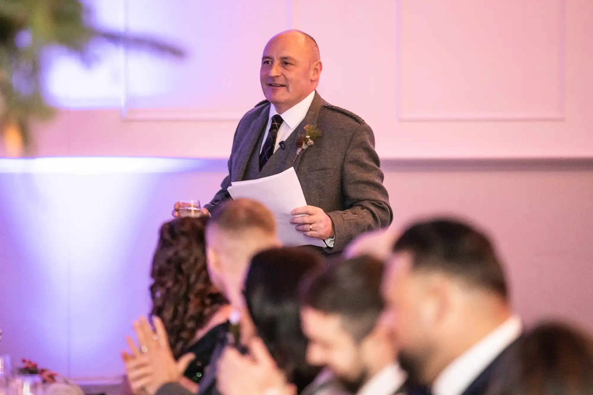 A man in a suit stands holding a piece of paper and a drink, speaking to seated guests at an indoor event.