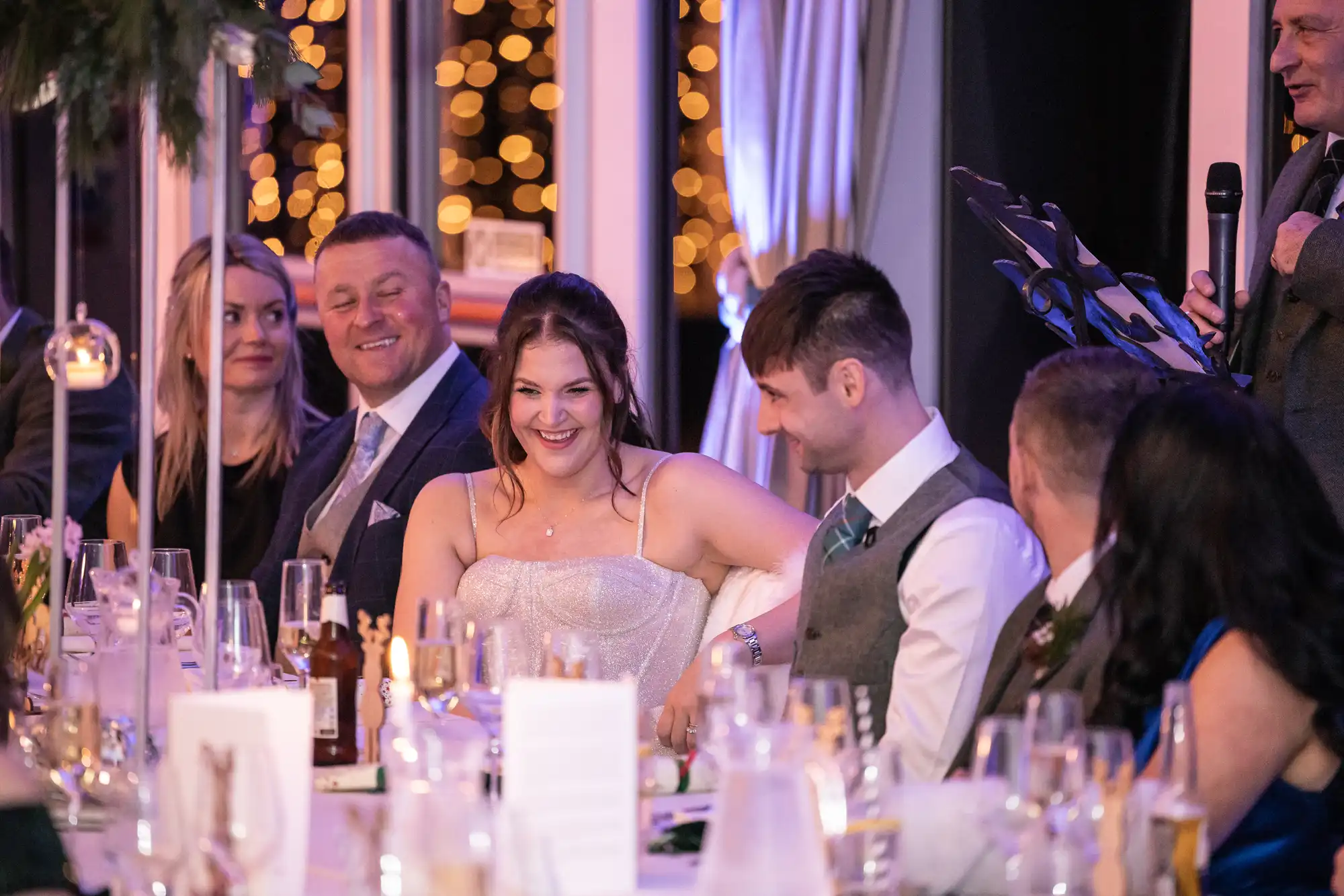 A group of people are seated at an elegantly decorated table, smiling and engaged in conversation. One man stands at a microphone, possibly giving a speech, with warm lighting illuminating the background.