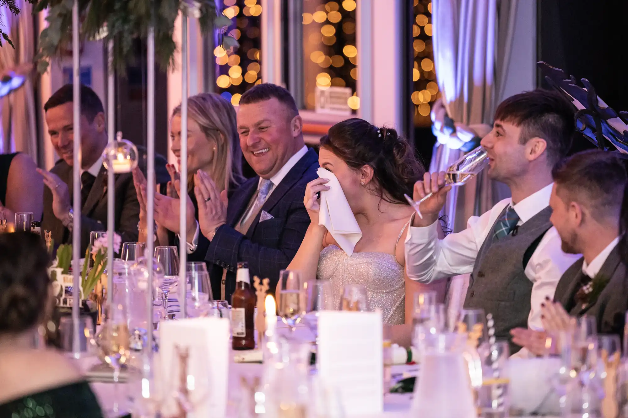 A bride wipes her face with a napkin while guests clapping and smiling are seated at a wedding reception table decorated with candles and lights.