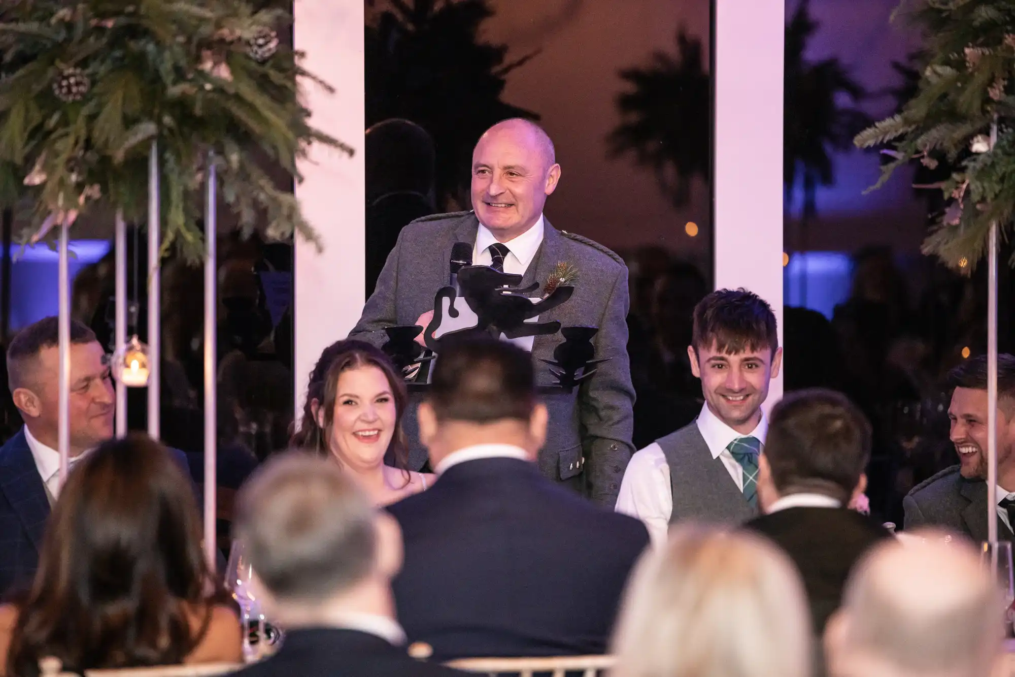 A man stands and speaks at a formal event with people seated around tables. Several attendees are smiling, and the room is decorated with hanging greenery.