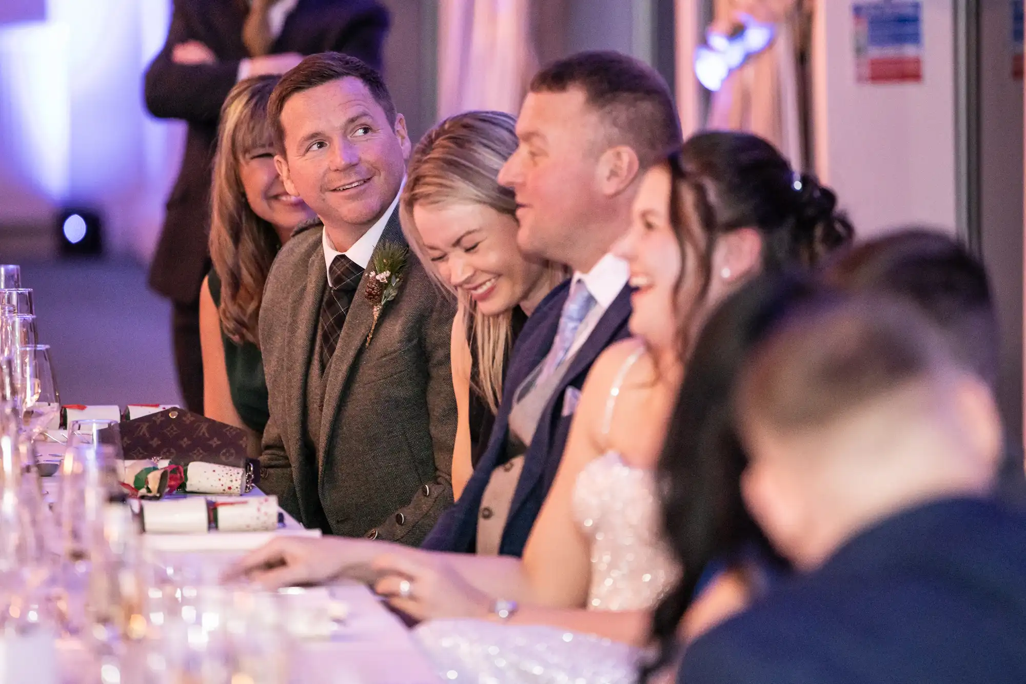 A group of people dressed in formal attire sit at a long table, smiling and conversing. Some have drinks, and one has a gift box in front of them. It appears to be a celebratory event.